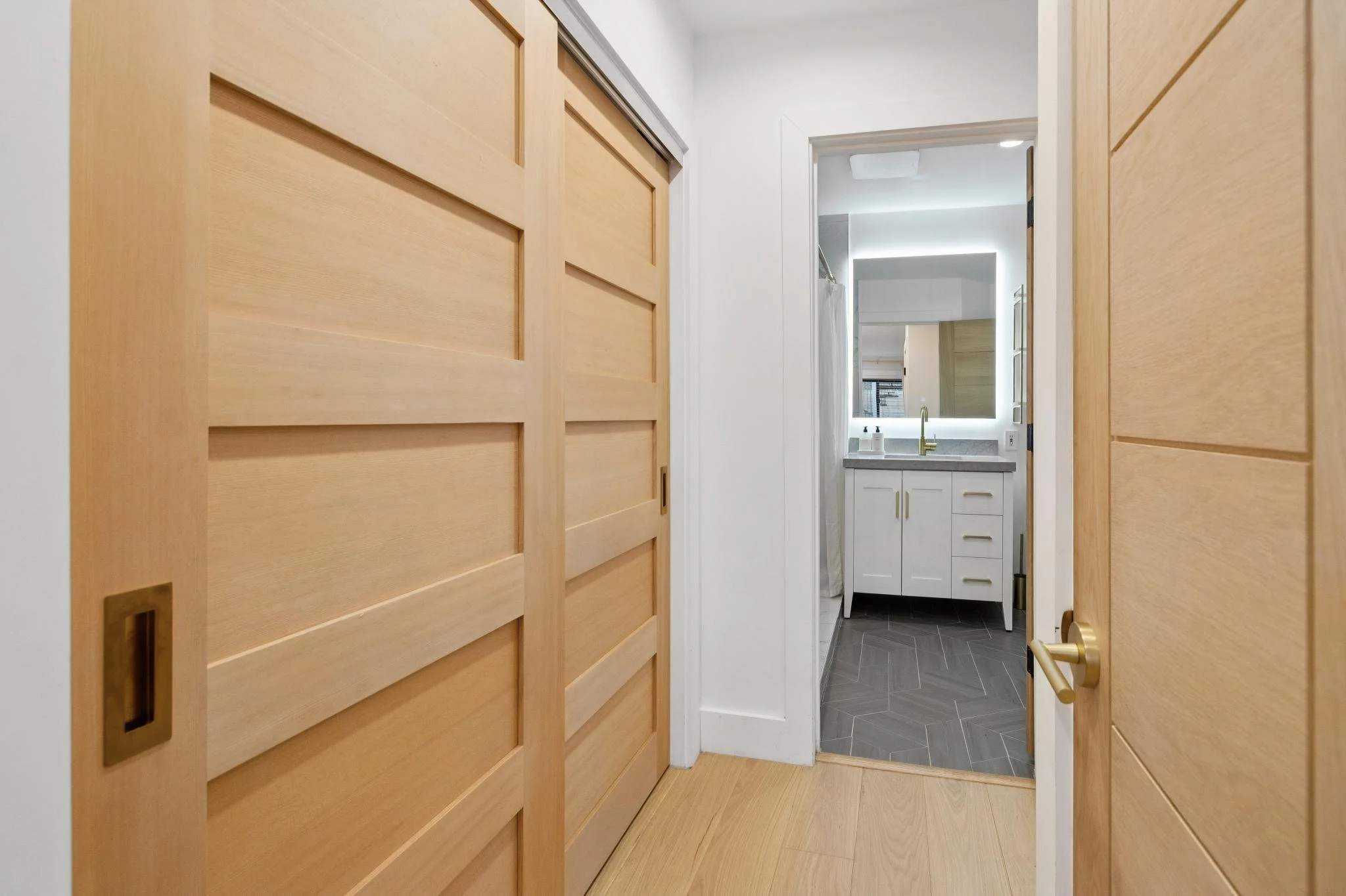 View of a small bathroom seen through wooden sliding doors, with a white vanity and a large mirror illuminated by built-in lighting.