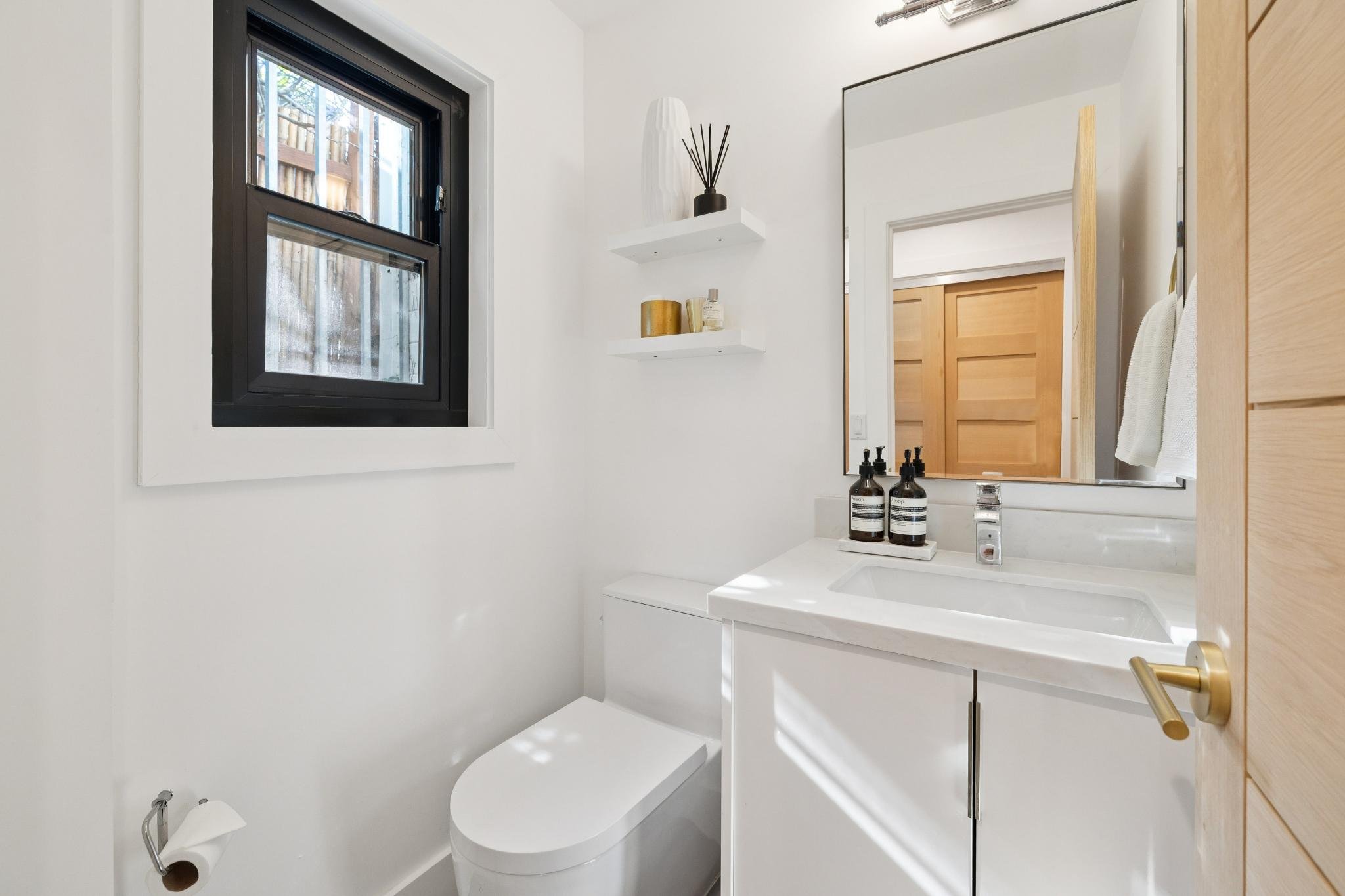 Small modern bathroom with a black-framed window, white walls, a white toilet, a white vanity with a sink, a large mirror, black soap dispensers, and minimal decor on shelves.