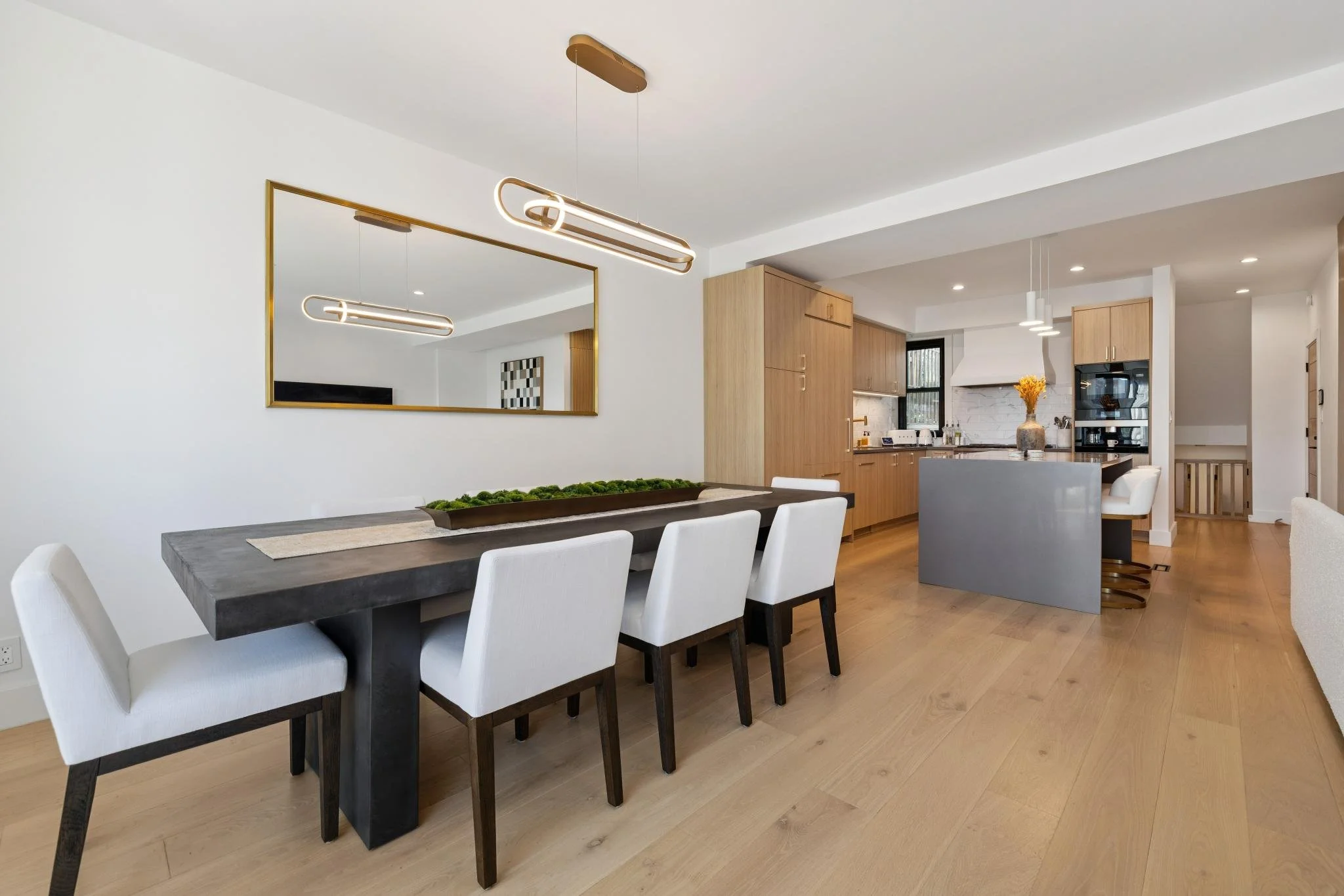 Modern dining room and kitchen with light wood flooring, white walls, rectangular mirror, black table, white chairs, and contemporary light fixtures.