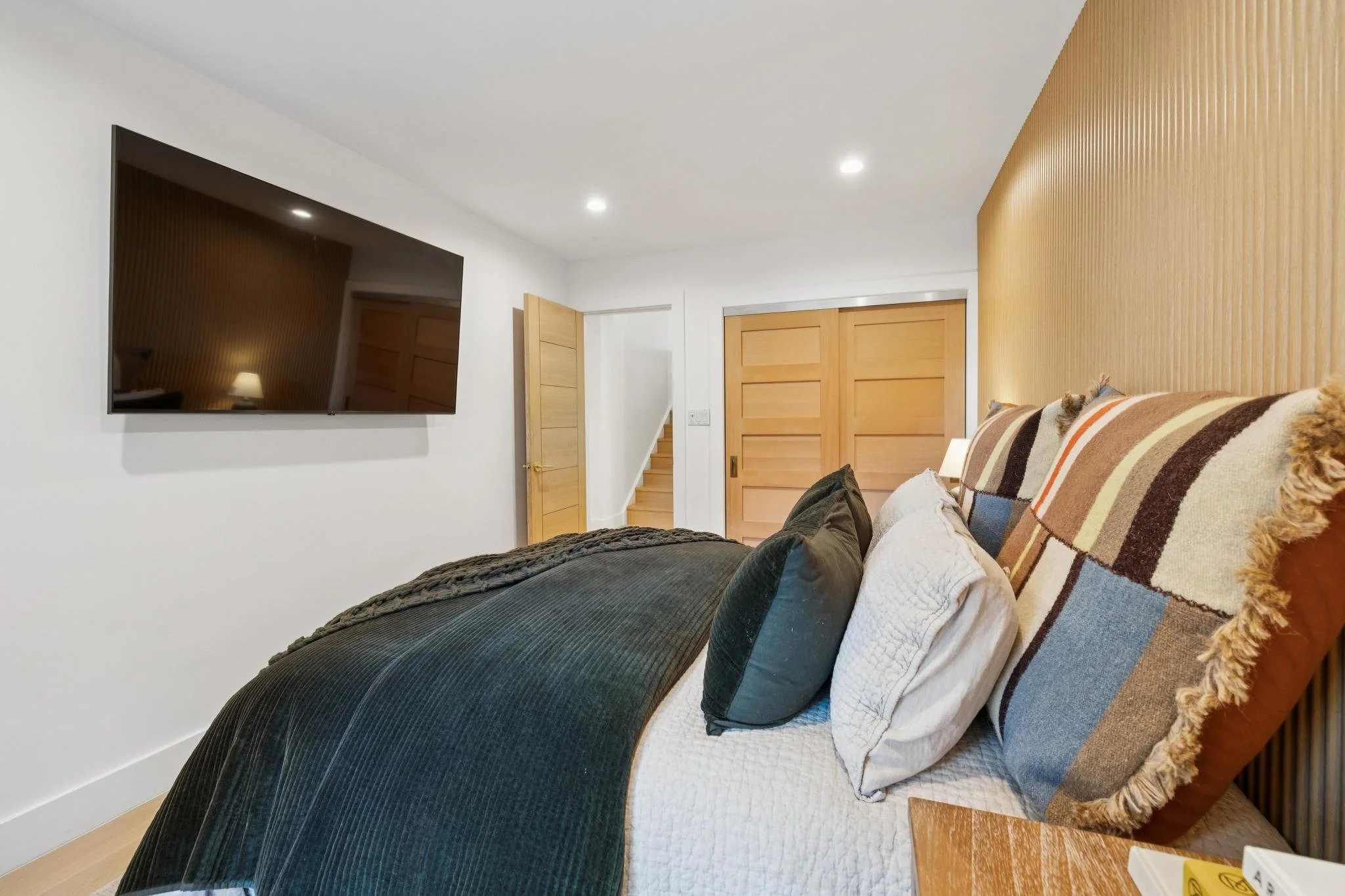 Modern bedroom with a large bed, plaid headboard, and dark bedding, wall-mounted TV, wooden sliding doors, and a small bedside table.