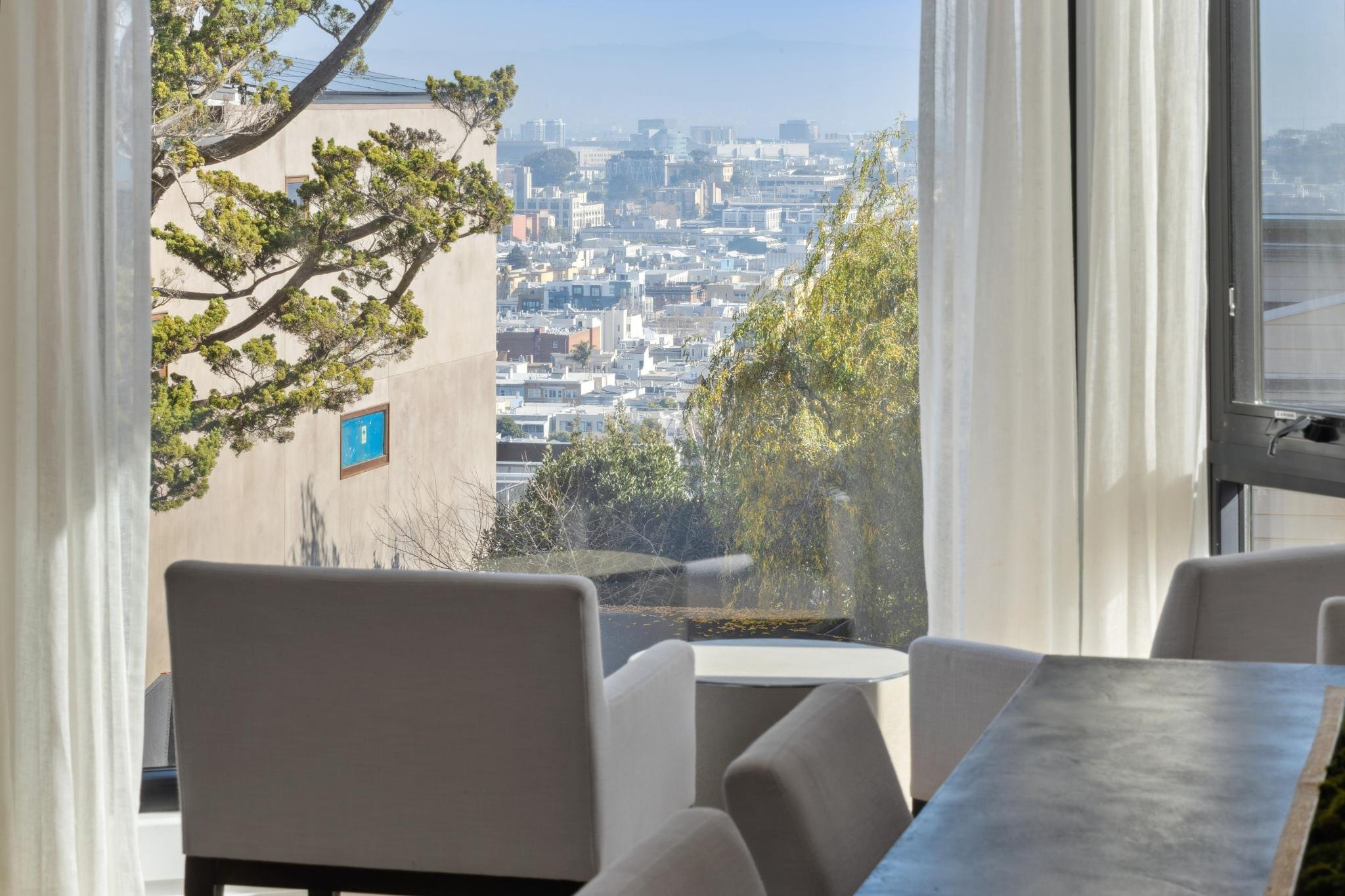 A bright hotel room with large window showing city view and trees outside; white chairs and a table inside.