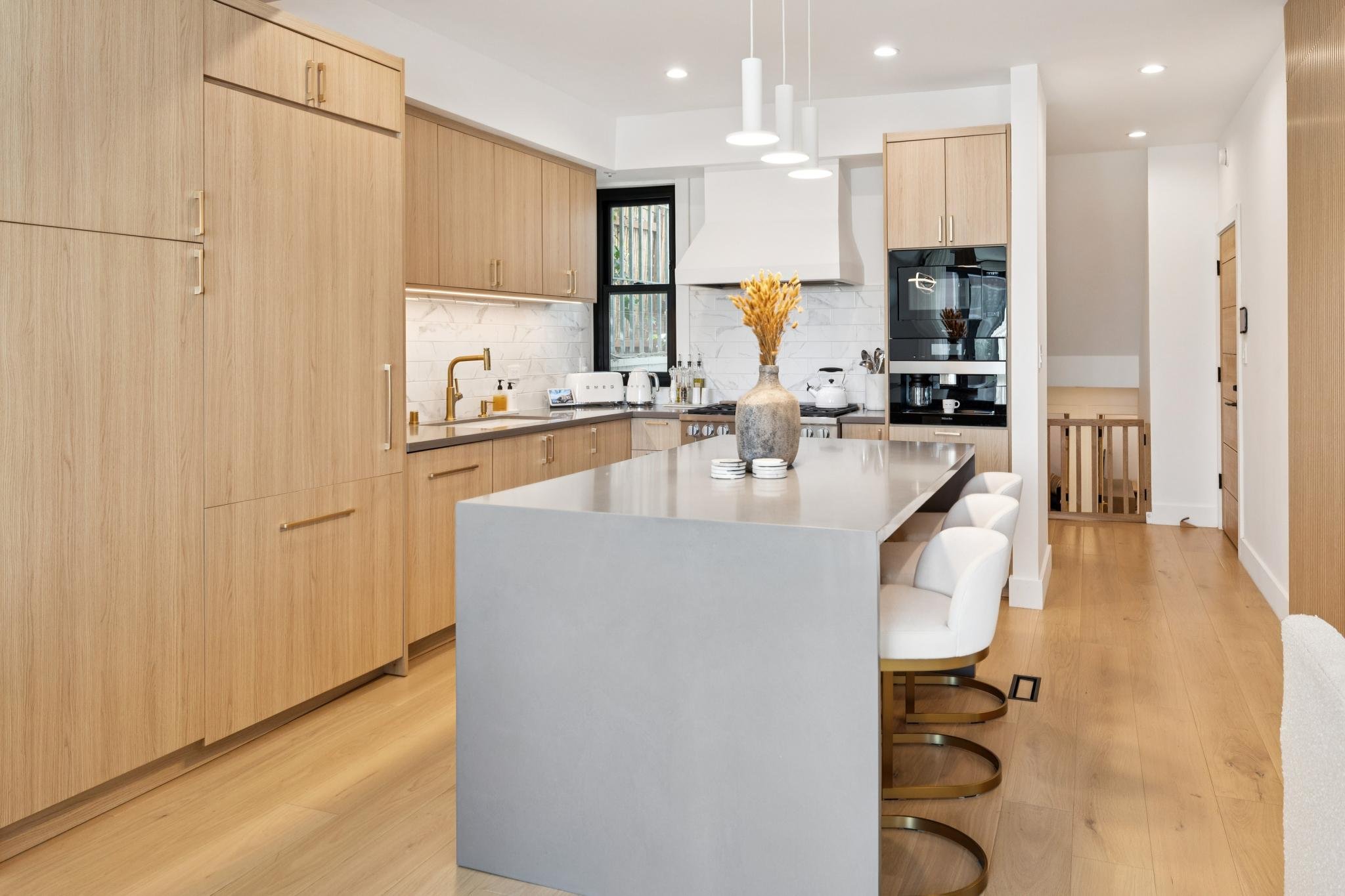 Modern kitchen with light wood cabinets, a large center island with a gray countertop, four white chairs with brass legs, and minimal decor including a large vase with dried flowers, set against white walls and hardwood floors.