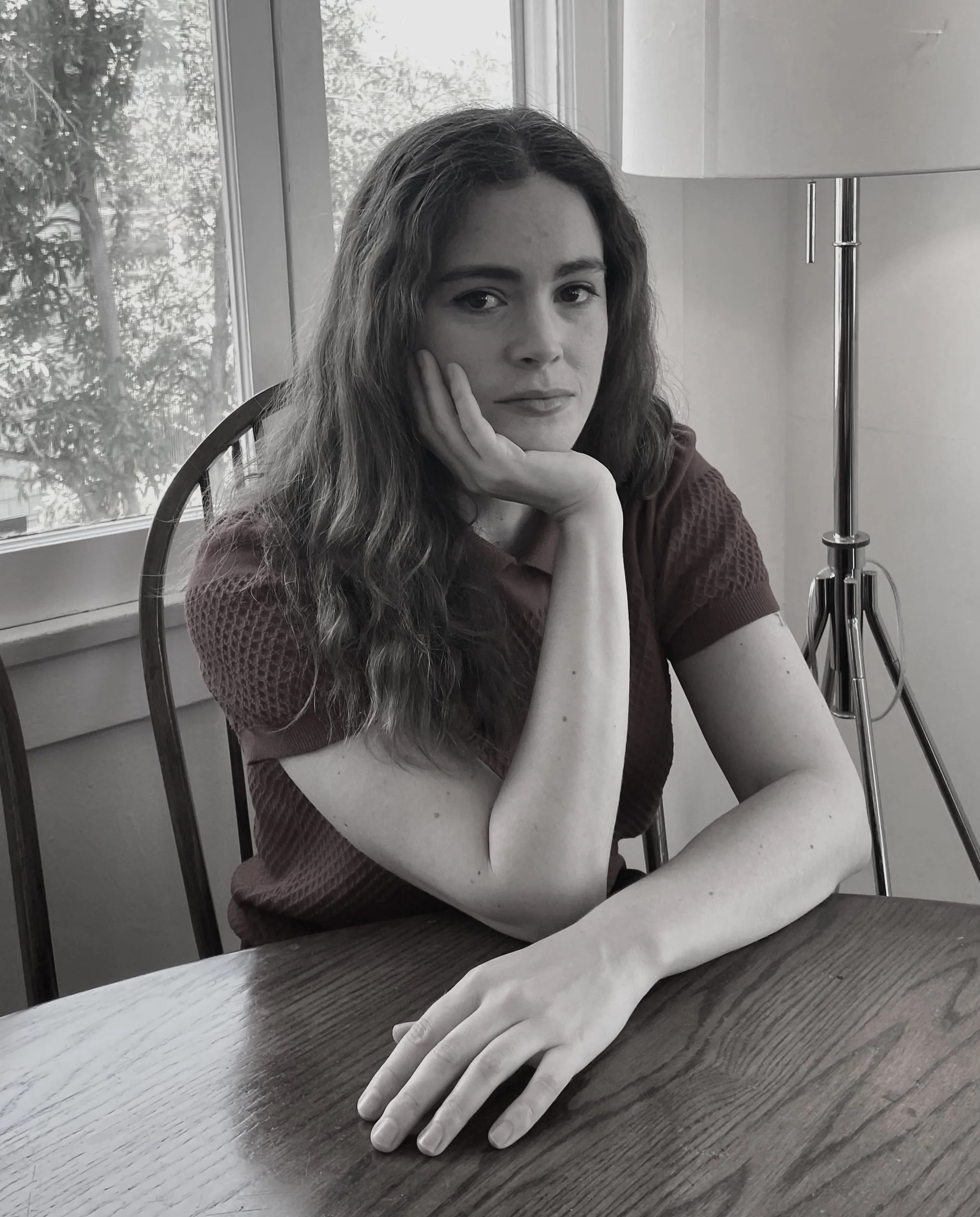 A young woman with wavy hair sitting at a wooden table near a window, resting her head on her hand, with a thoughtful expression.
