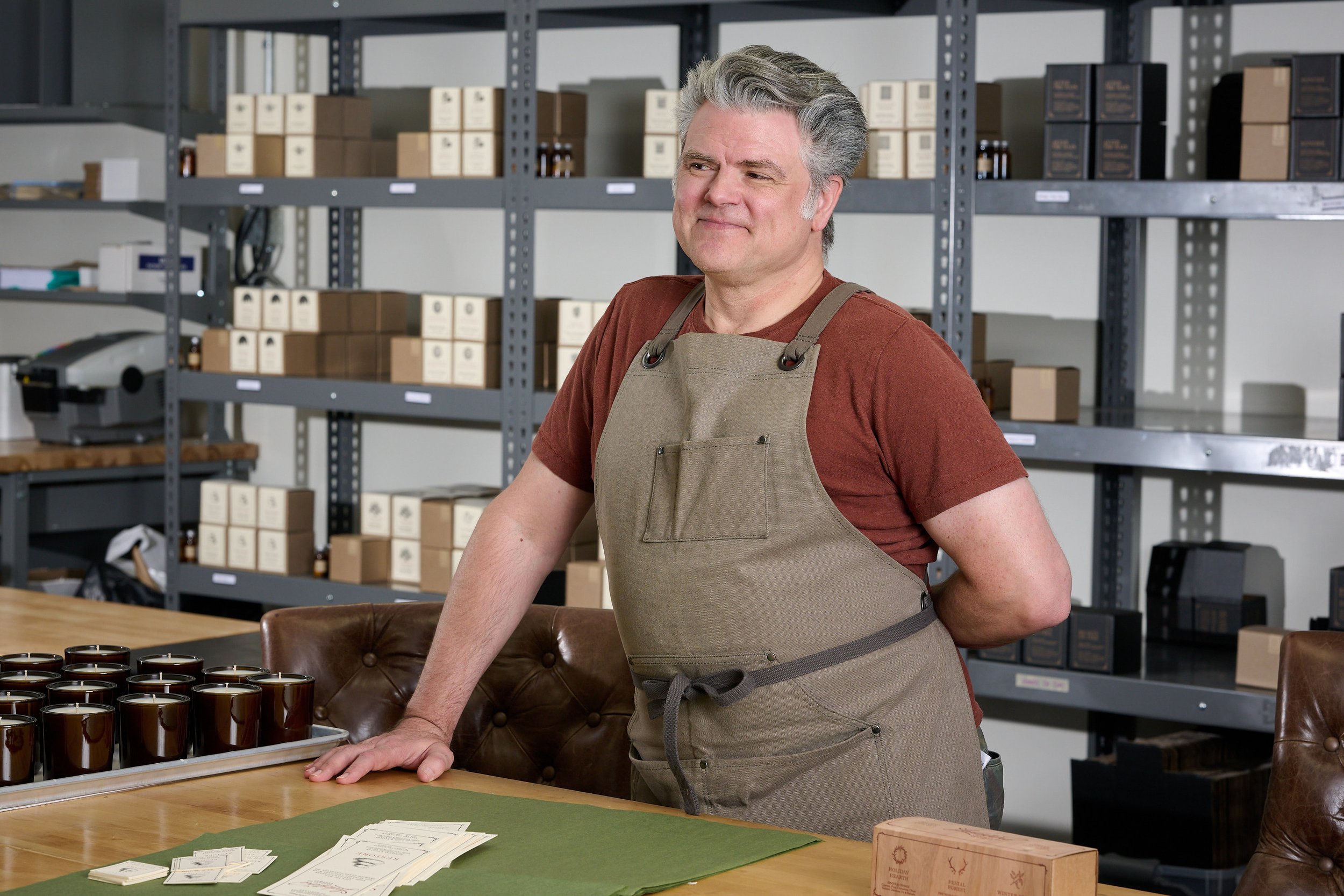 A middle-aged man with gray hair in a brown T-shirt and a beige apron, standing behind a wooden table in a storage room or workshop. The table has several brown coffee mugs and some papers on it. Behind him are metal shelves stocked with cardboard bo