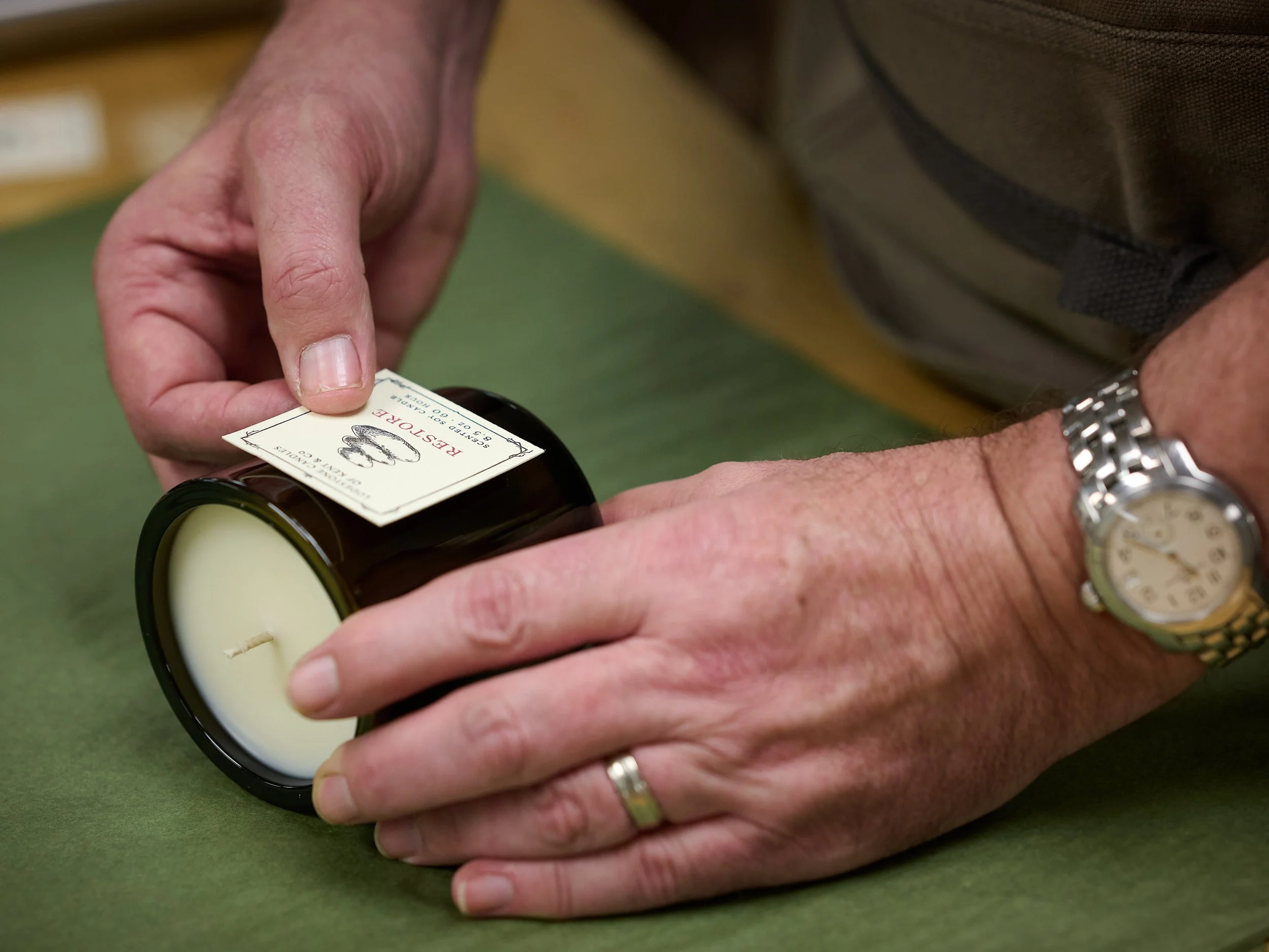 A person with a silver wristwatch and wedding band is holding a scented candle in a glass jar. The person is peeling off a label from the jar, which has a white candle inside.
