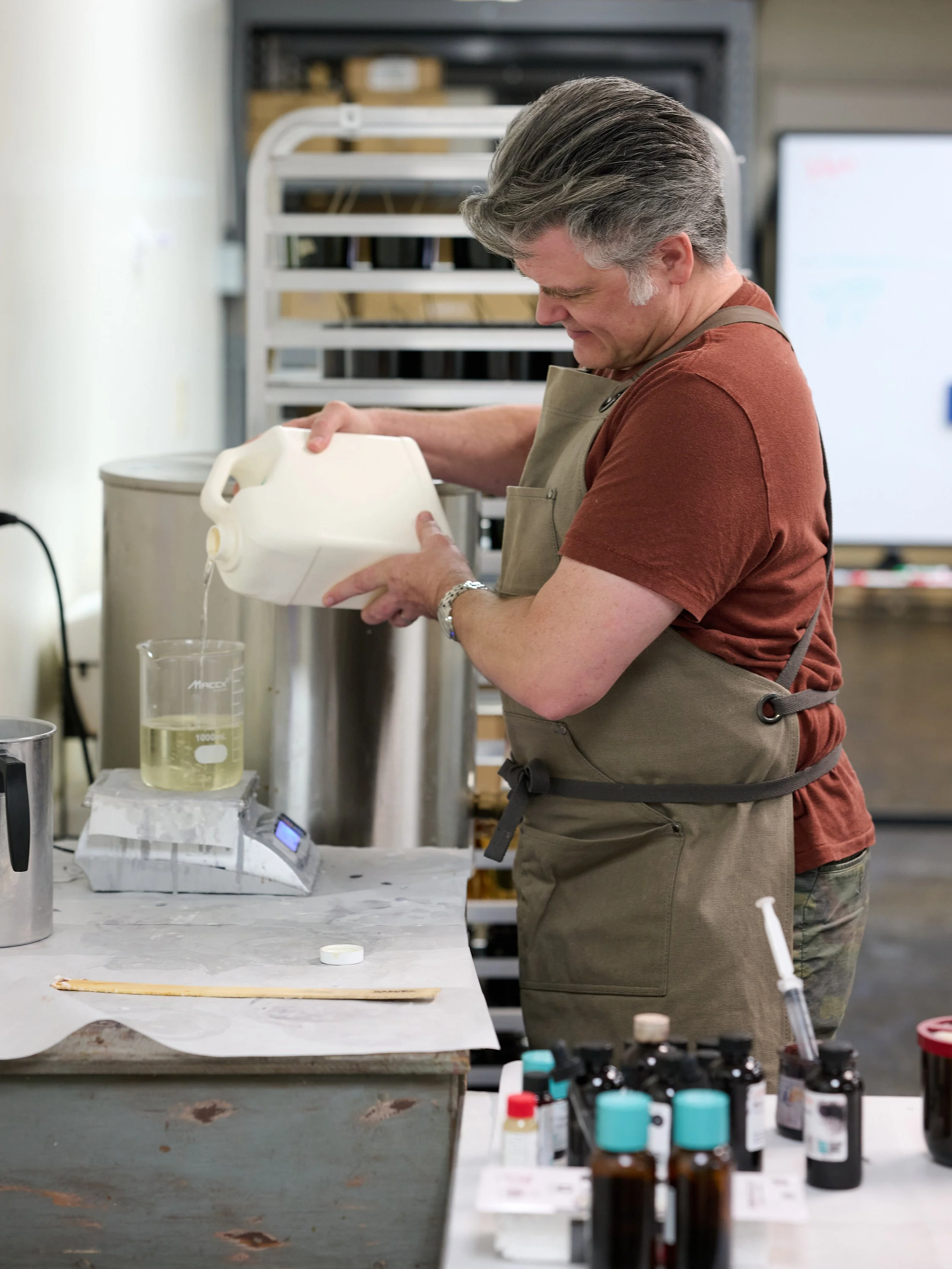A man wearing a beige apron pouring a liquid into a beaker in a laboratory or workshop setting, with various bottles and equipment on the table.