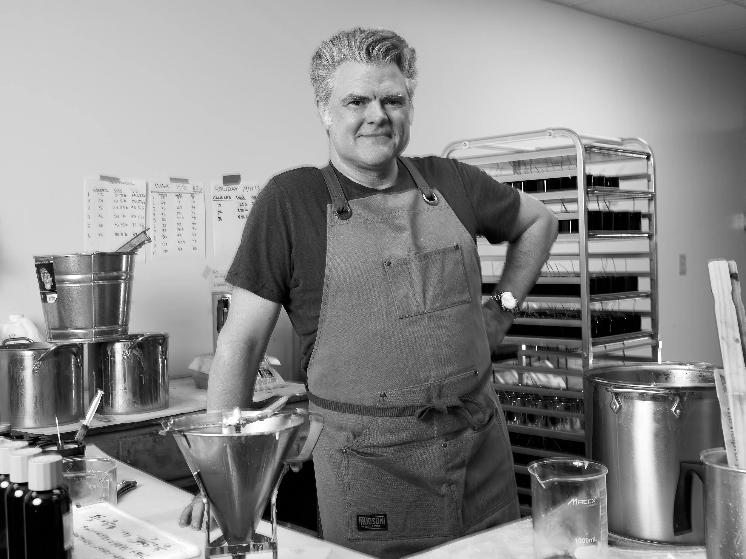 A man in an apron standing in a laboratory or workshop surrounded by equipment, with a rack of trays and papers on the wall behind him.