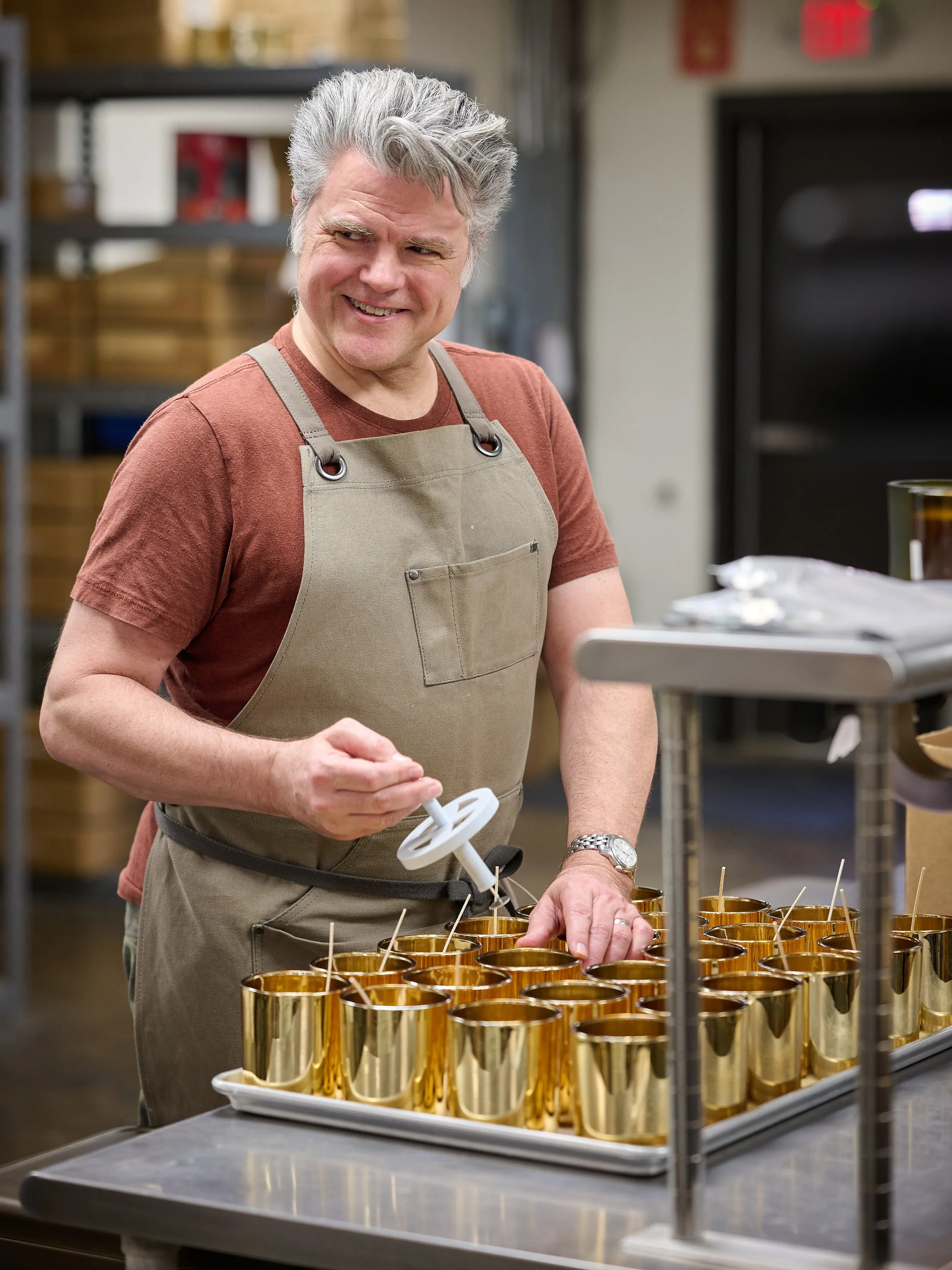 A man with gray hair wearing a brown t-shirt and tan apron is working in a kitchen, filling gold-colored cups with a syringe-like tool.
