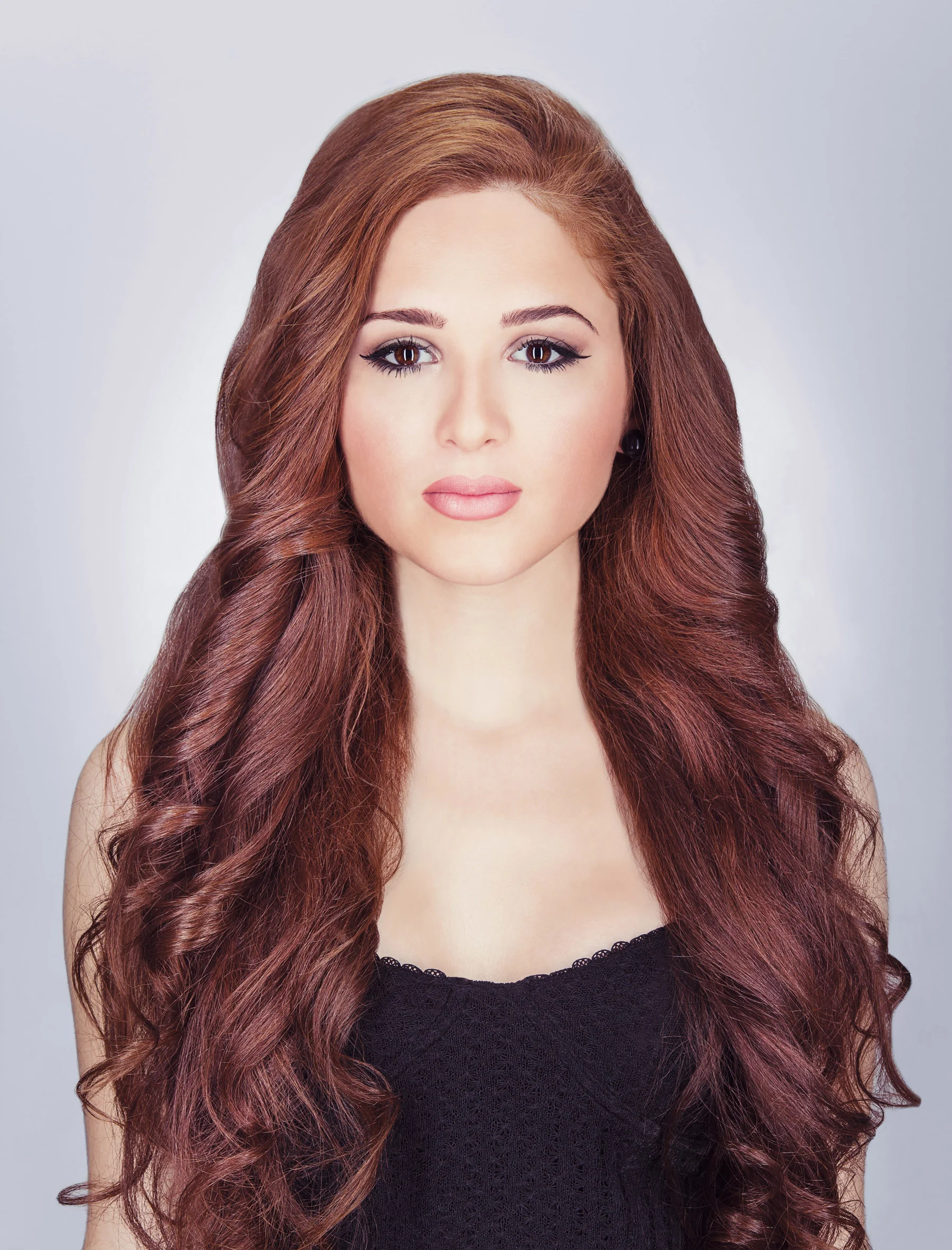 Portrait of a young woman with long, wavy auburn hair, wearing a black sleeveless top, standing against a plain light gray background.