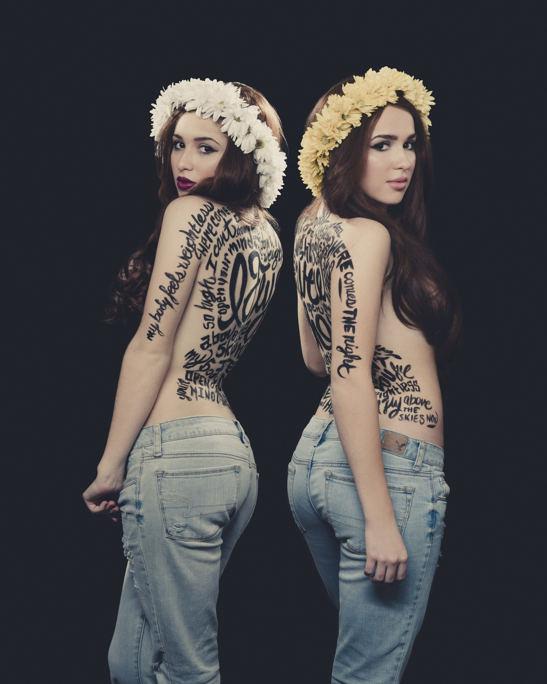 Two women with long brown hair wearing flower crowns, one white and one yellow, stand with their backs to each other against a black background. They are topless with black writing tattoos covering their backs and sides, wearing light blue jeans.
