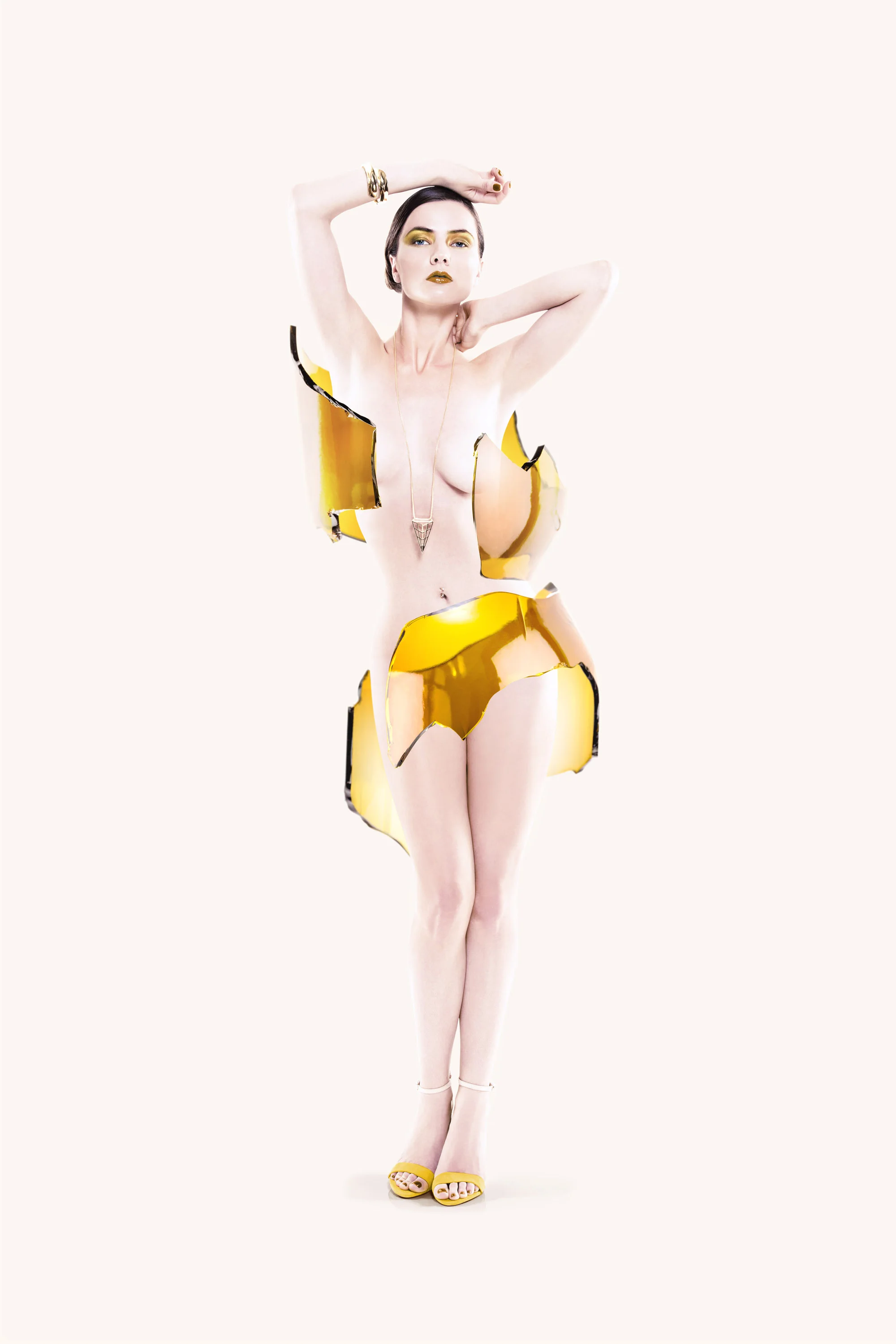 A woman with short dark hair and dark lipstick, wearing yellow eyeshadow, poses with her arms above her head. She is dressed in a highly stylized, conceptual outfit made of reflective, metallic gold and black pieces resembling broken fragments, expos
