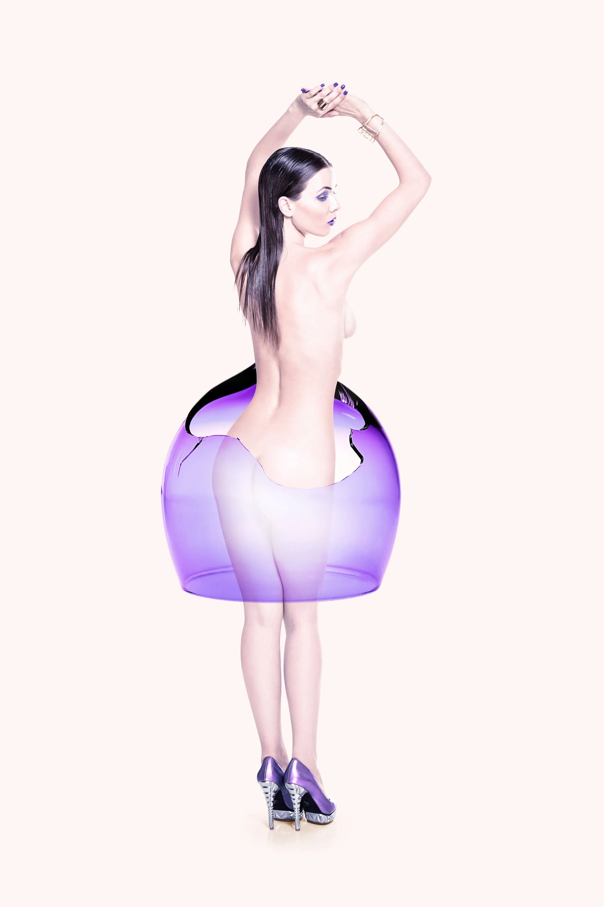A woman with long dark hair is standing inside a large purple glass bowl that appears to be broken. She is topless, wearing purple high-heeled shoes, and has her arms raised above her head, holding her hands together.