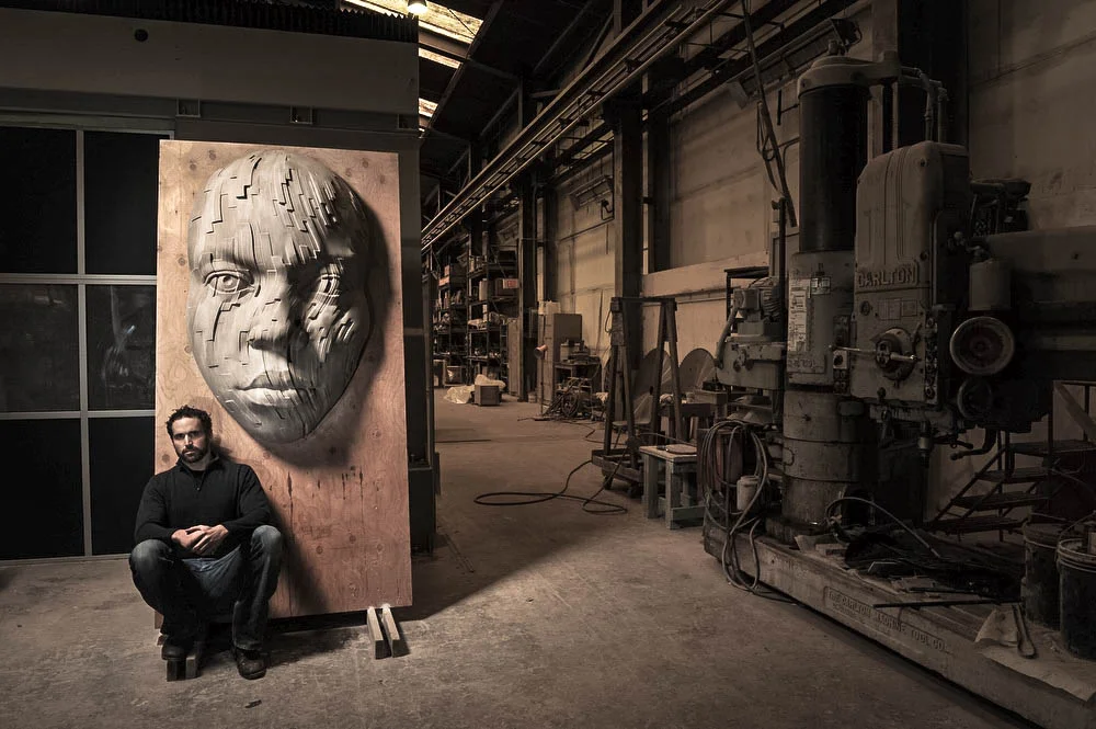 An artist in a black shirt sitting on a stool in an industrial workshop next to a large wooden panel with a 3D face sculpture on it, featuring an abstract, pixelated design.