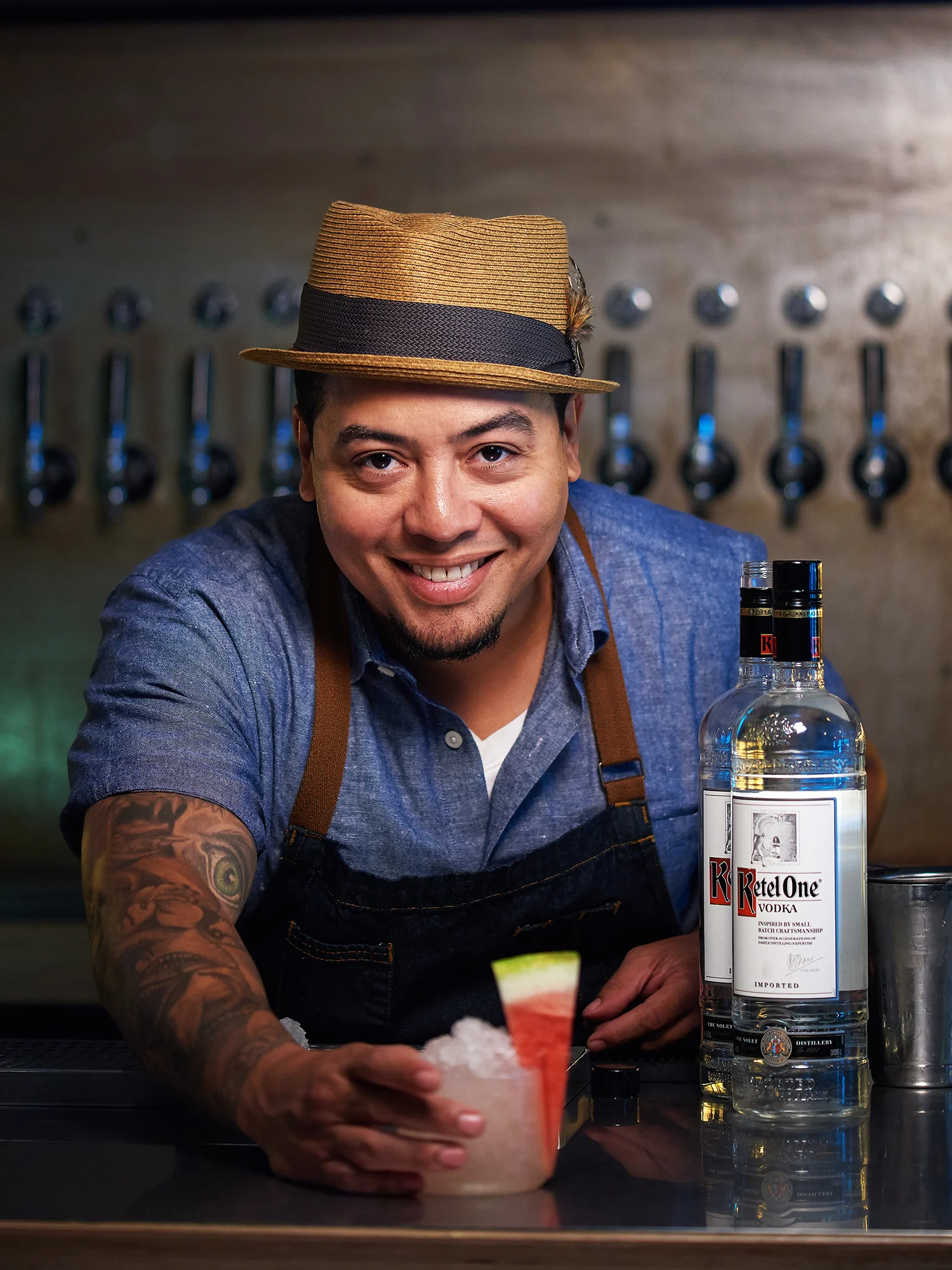 A smiling bartender in a blue shirt, wearing a straw hat, is holding a drink with a watermelon garnish, sitting at a bar with bottles of vodka behind him.