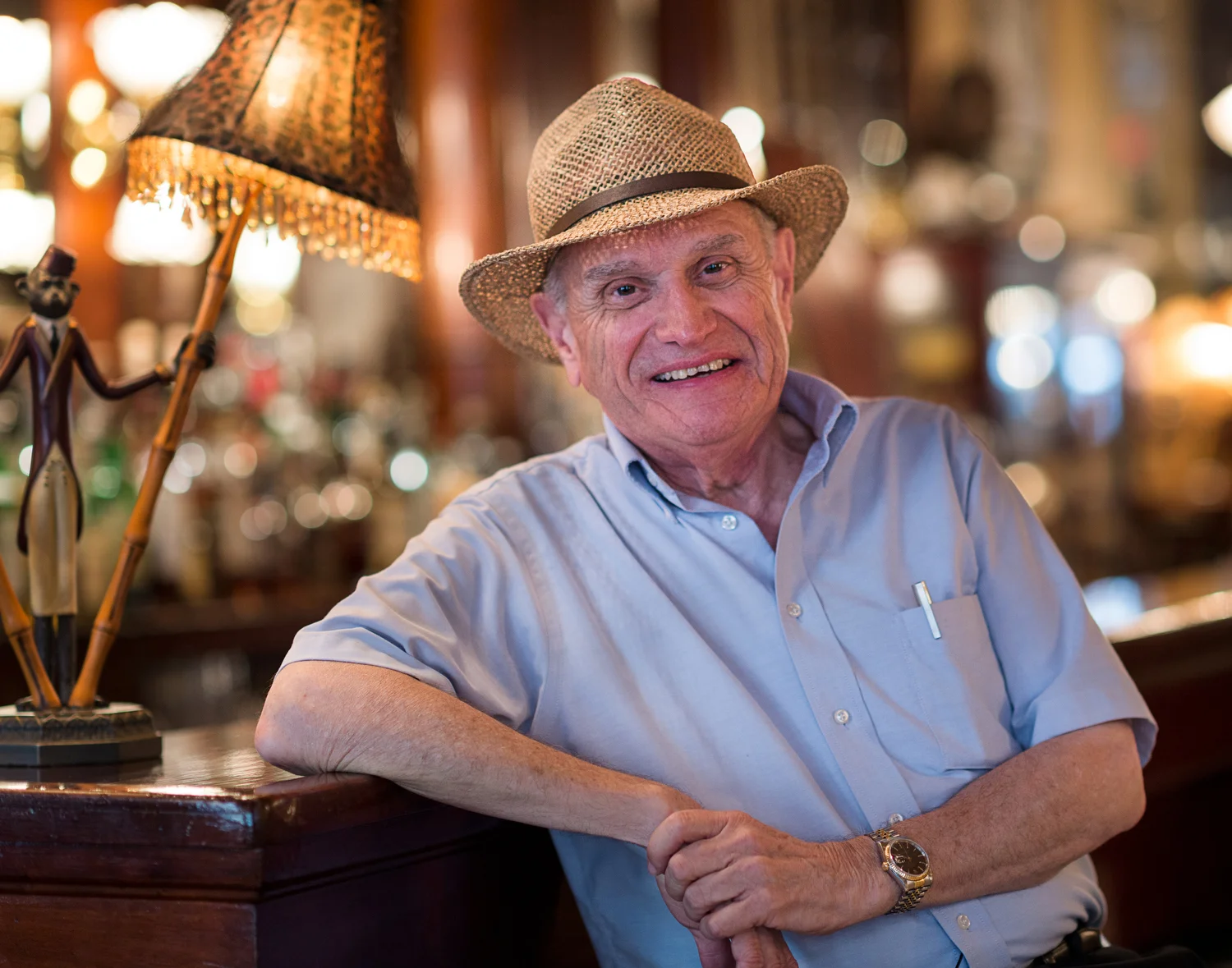 An elderly man wearing a straw hat, light blue button-up shirt, and gold watch, smiling and sitting at a bar counter with a vintage lamp and blurred background.