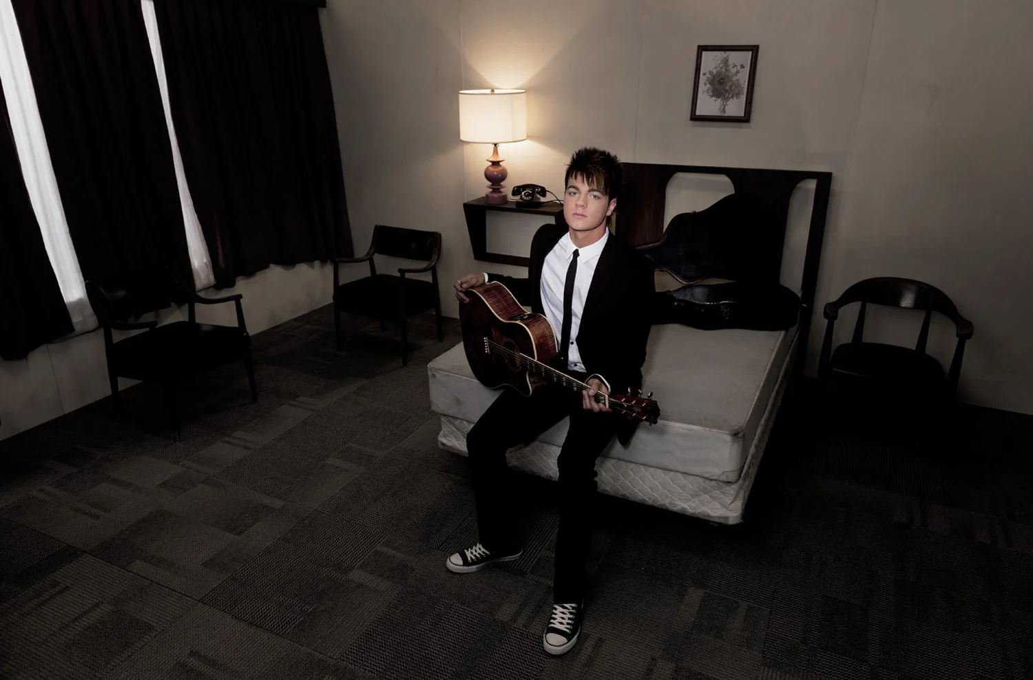 A young man in a black suit, white dress shirt, and black tie sitting on the edge of a mattress in a dimly lit hotel room, holding an acoustic guitar, with a serious expression on his face.