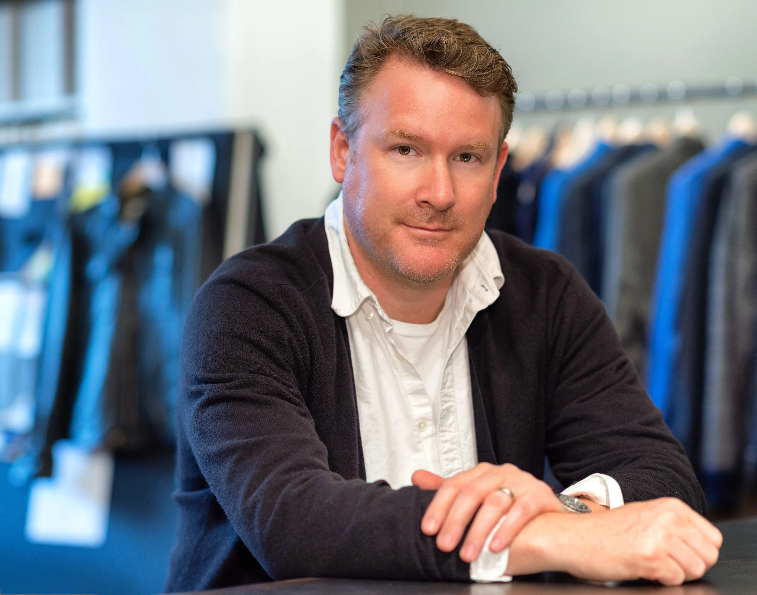Man with light brown hair, wearing a white shirt under a dark cardigan, sitting at a desk in a clothing store with racks of jeans in the background.