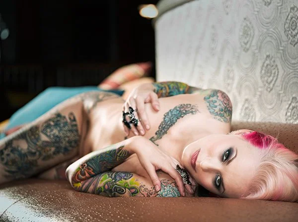 Tattooed woman with pink hair lying on her side on a bed, wearing jewelry and colored pants.