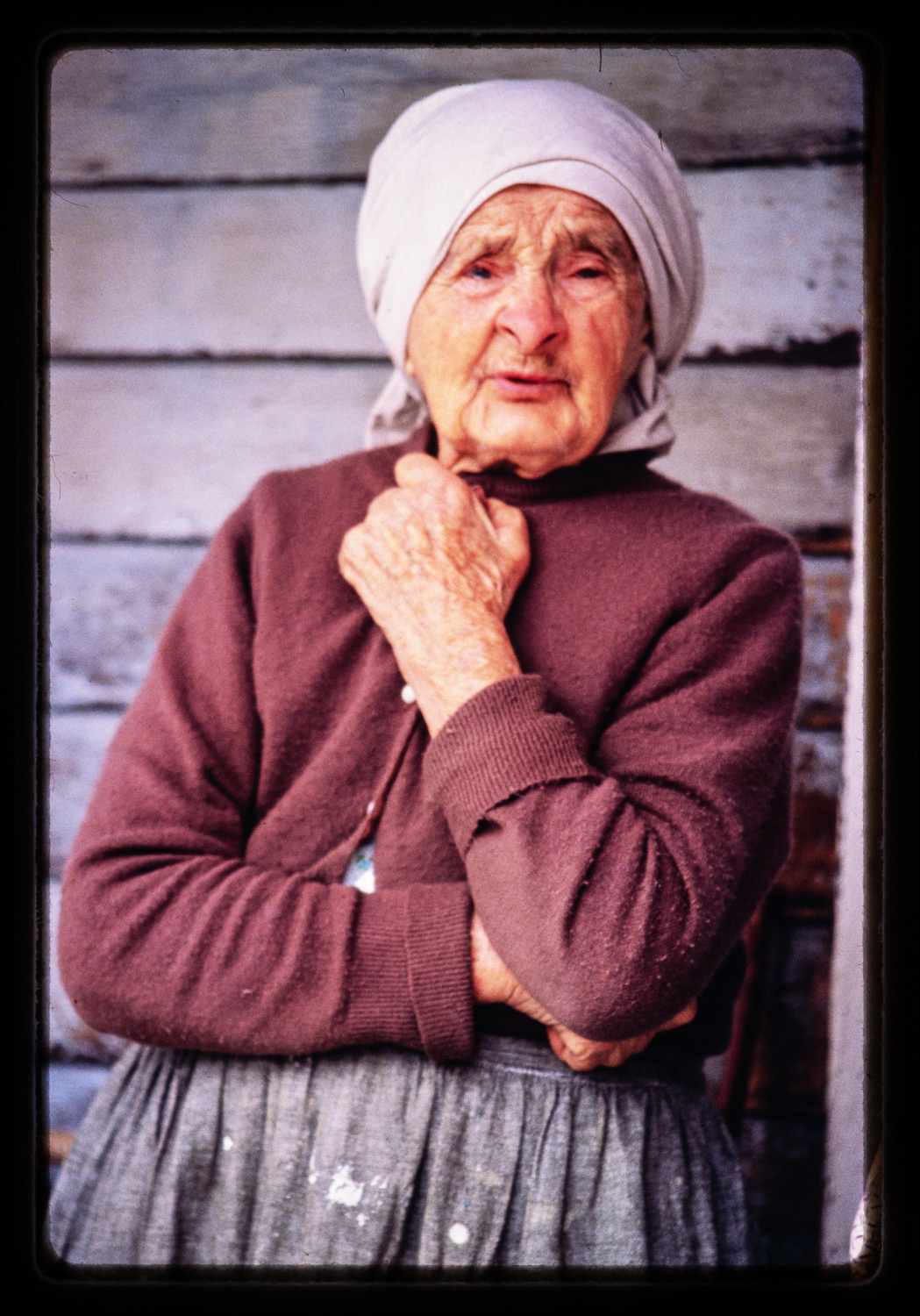 An elderly woman wearing a white headscarf, a brown sweater, and a gray skirt, standing in front of a wooden wall.