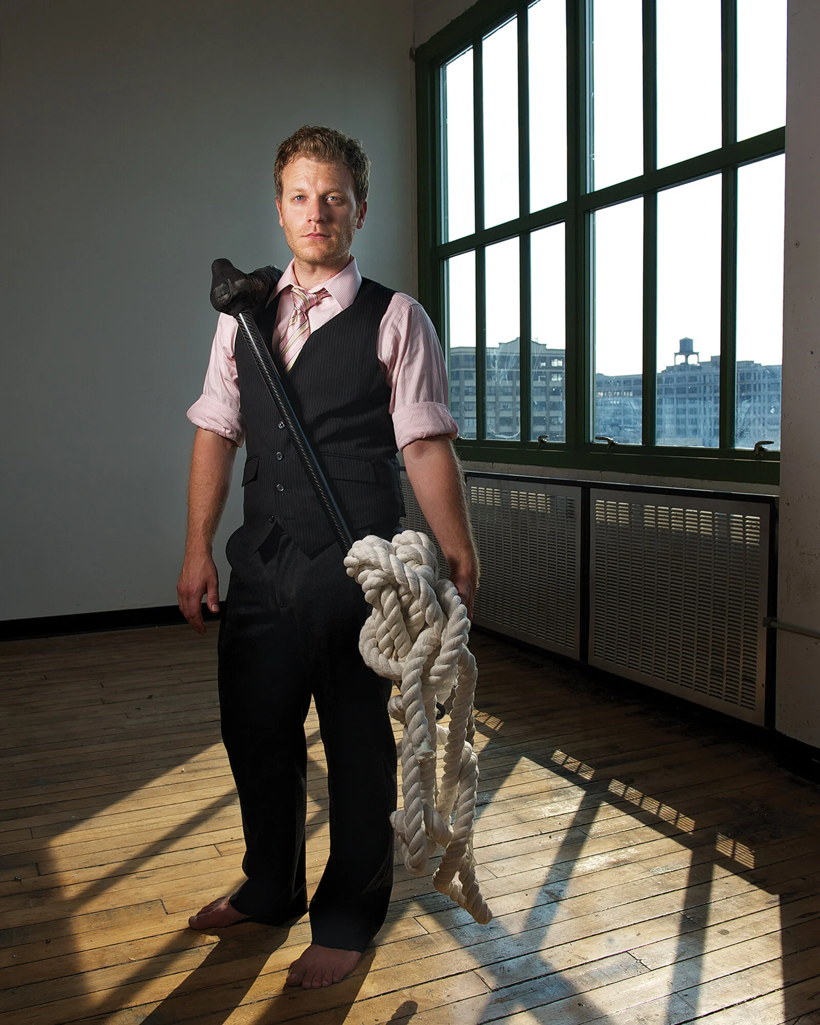 A man in business attire holding a large rope, standing barefoot in a room with wooden floors and large windows, with a cityscape visible outside.