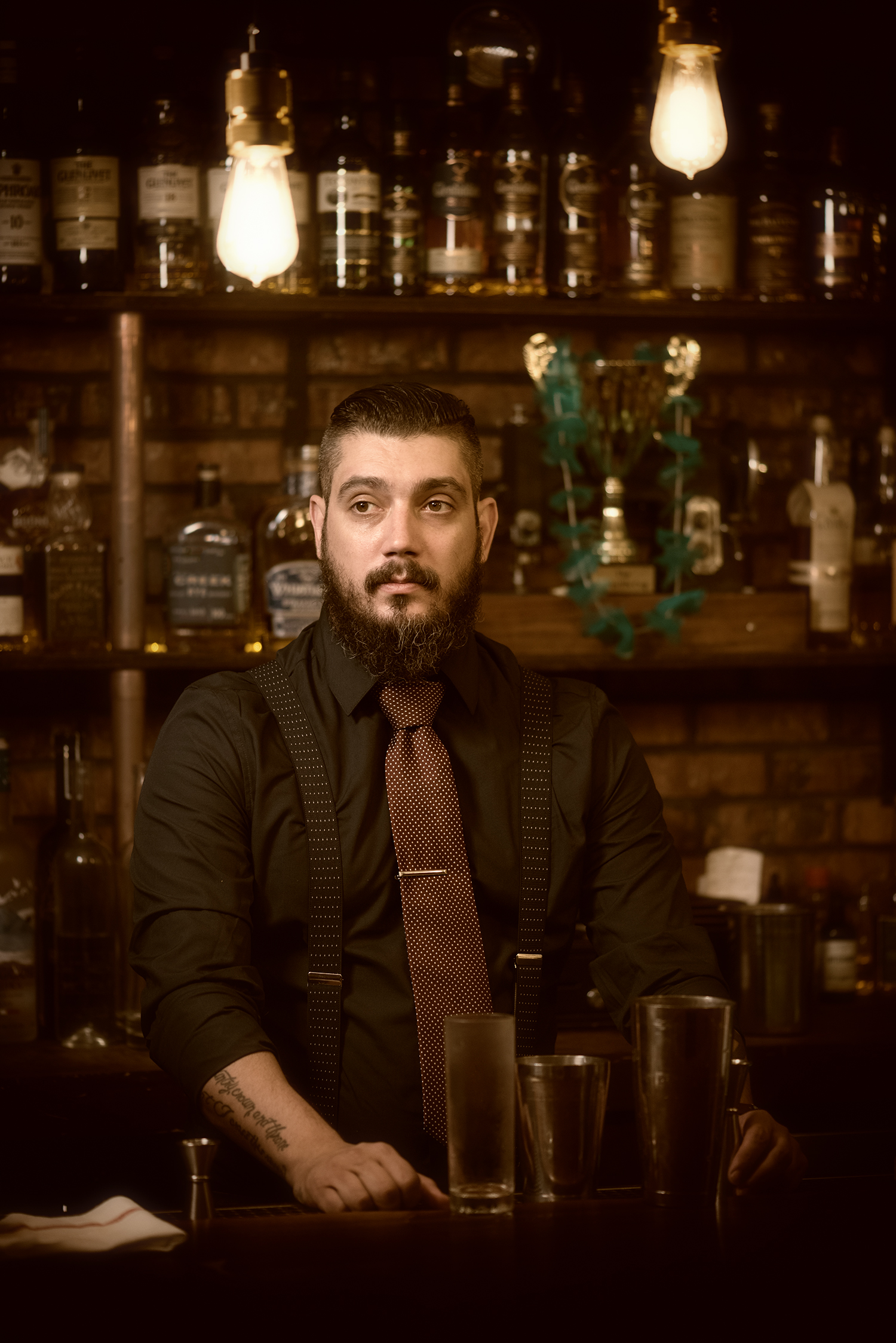 Bartender with a beard and short hair standing behind a bar with glasses, bottles, and dim warm lighting in the background.