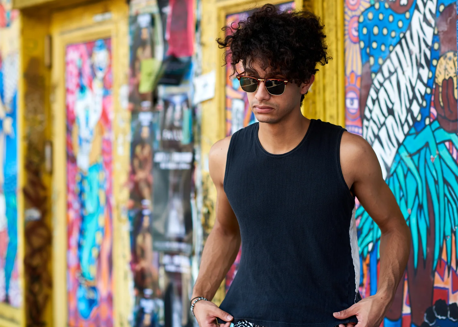 A young man with curly hair wearing sunglasses and a sleeveless black shirt walks along a colorful wall with graffiti art.