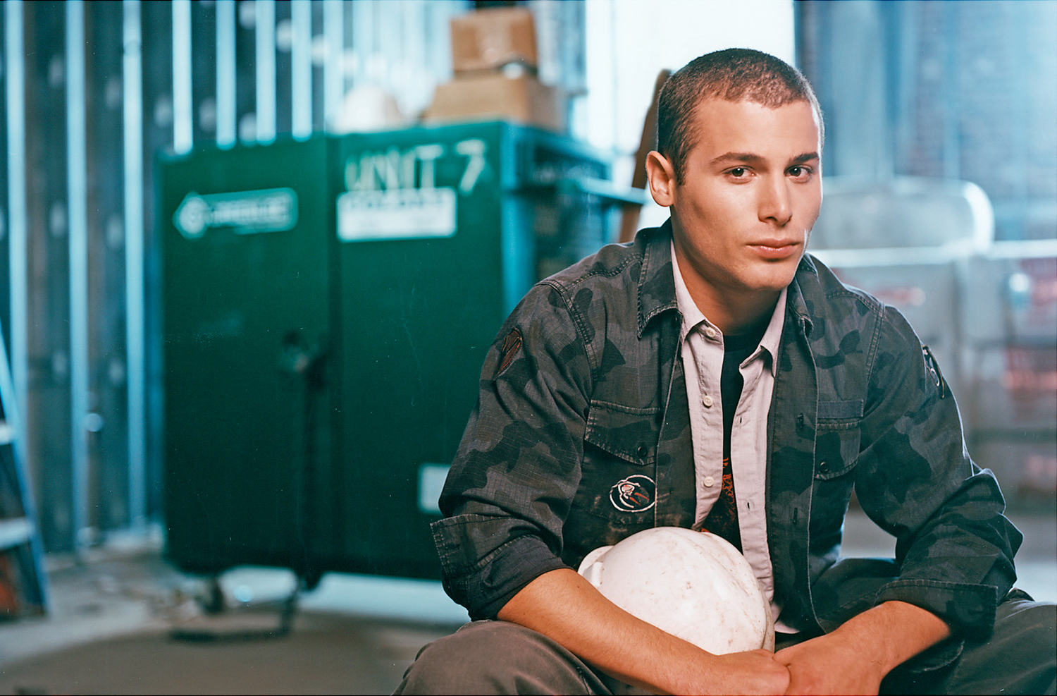 A young man with short hair, wearing a dark camouflage jacket over a light shirt, sits holding a white bowling ball in his lap inside a bowling alley. The background features a green storage cabinet and a blurred bowling lane.