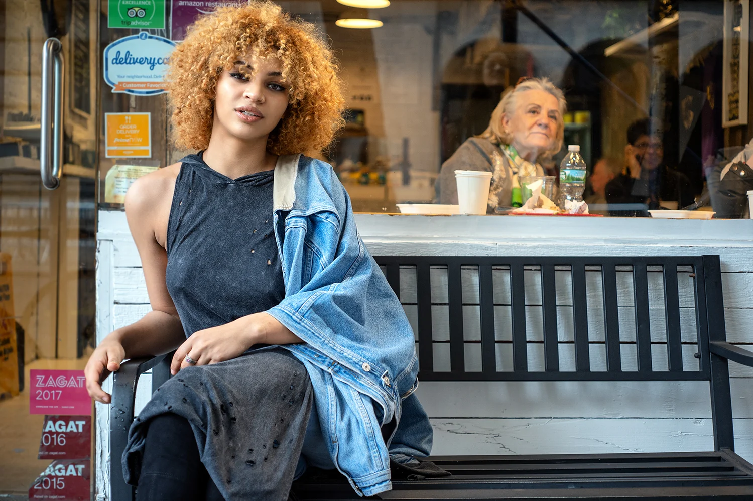 A young woman with curly blonde hair sitting on a black bench outside a restaurant, looking at the camera, with an older woman seated inside visible through the window behind her.