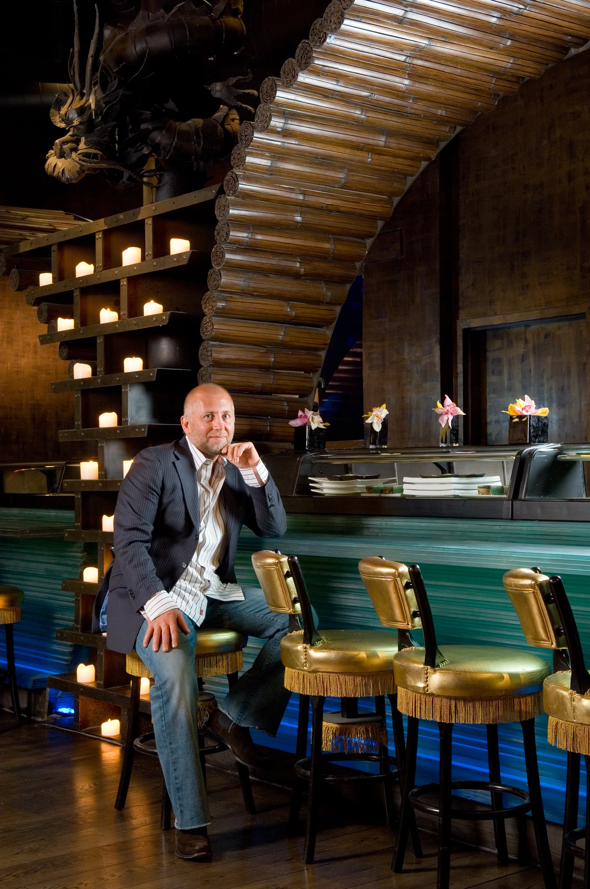 A man in a suit sitting on a gold-colored barstool at a bar with a modern, sleek interior. The background includes a staircase made of bamboo and a wall decorated with small candles and pink flowers, creating a warm and stylish ambiance.