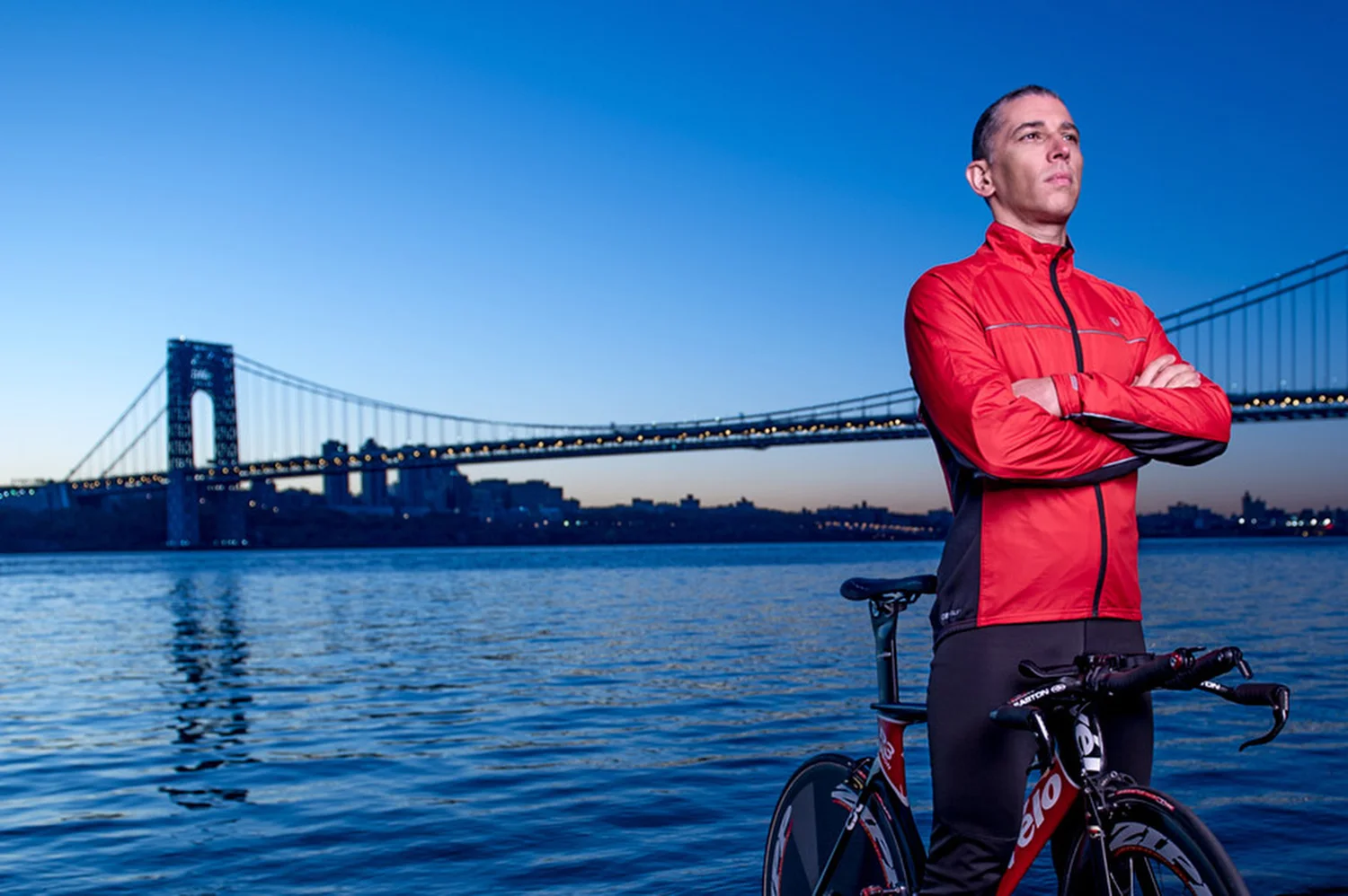 A man in a red jacket and black pants standing next to a bicycle by the water with a city bridge in the background at dusk.