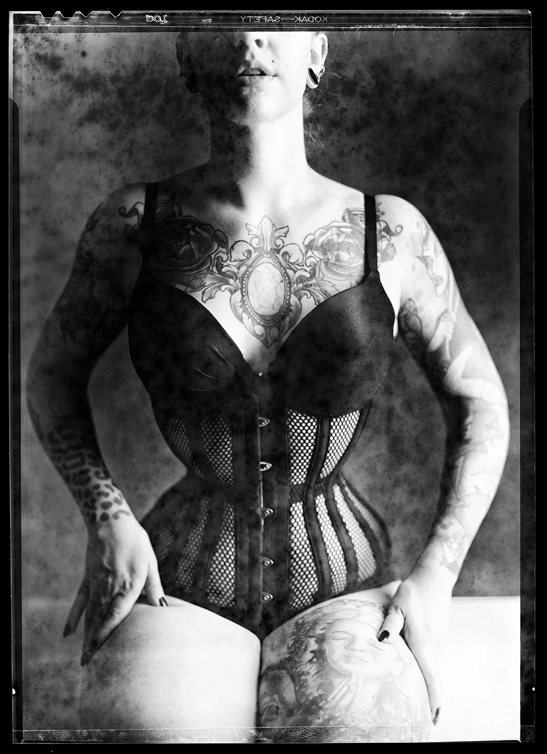 Black and white photograph of a tattooed woman wearing a black corset and mesh top, with tattoos on her arms, chest, and thigh, holding a tattooed globe in her lap.