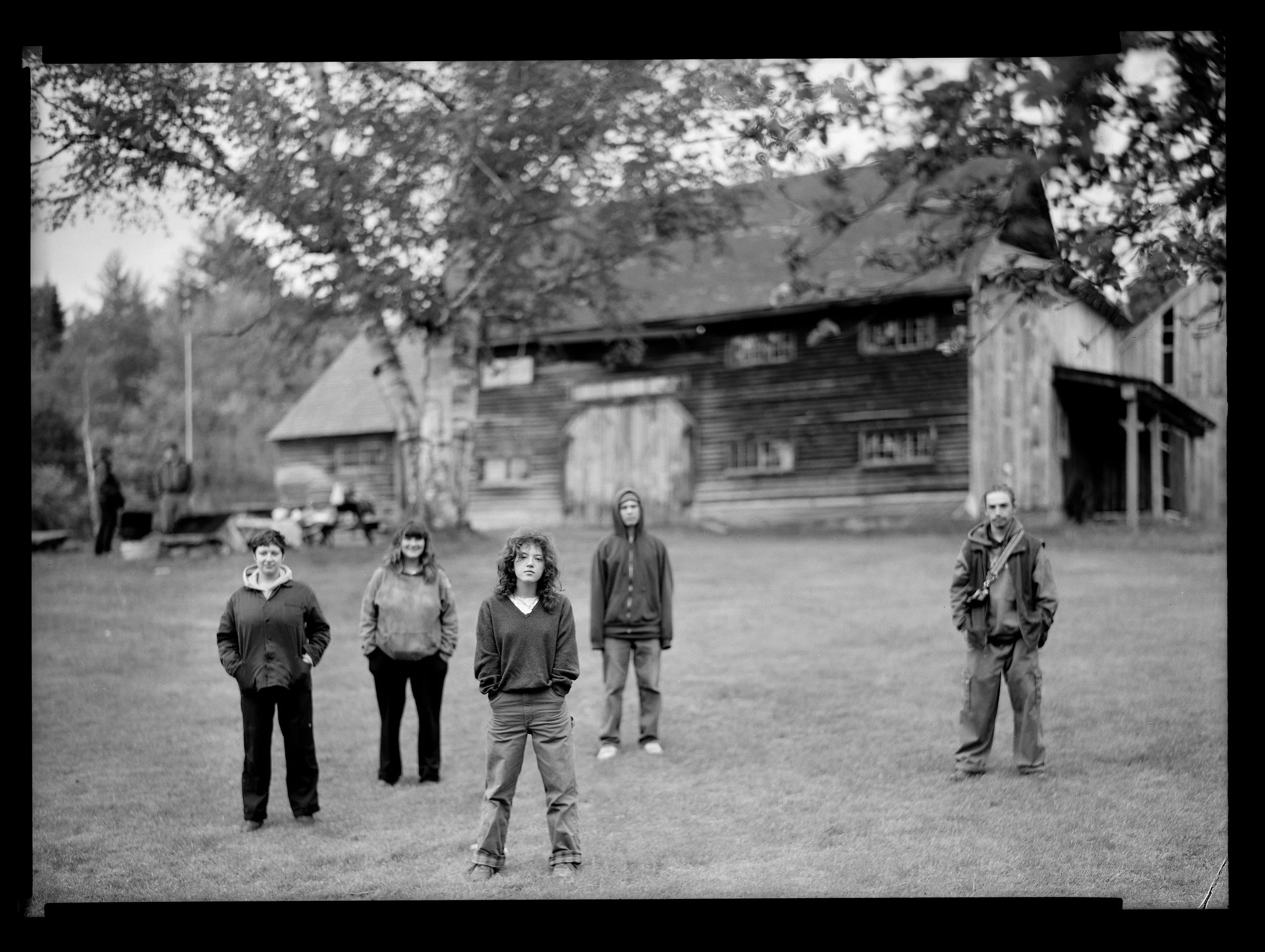 A black and white photo of a group of five people standing on a grassy area in front of a barn with a wooden exterior. The group appears to be posing for the photo, with three women and two men. There are trees around the barn and a rustic outdoor se