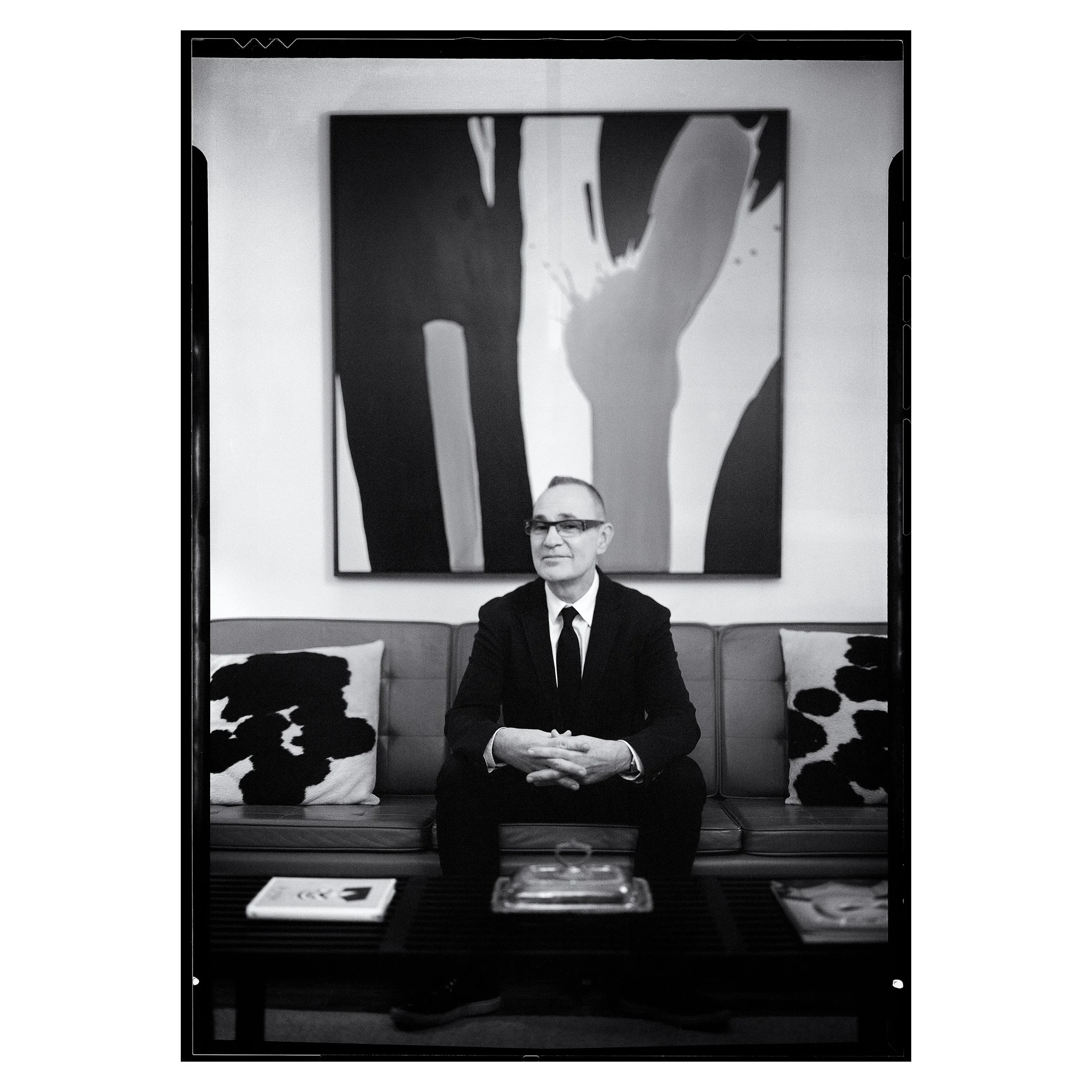A man in a suit and glasses seated on a sofa in a modern, art-filled room.