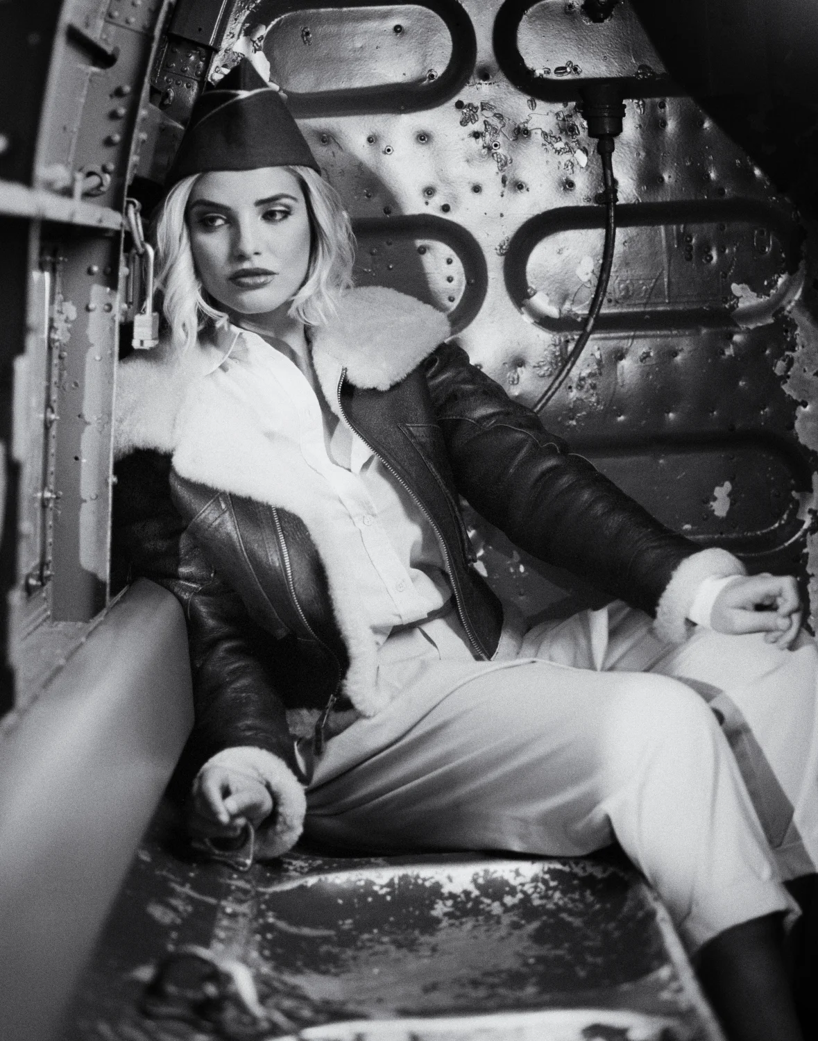 A woman with blonde hair in a vintage pilot jacket and pants, sitting inside the cockpit of an aircraft, in black and white.