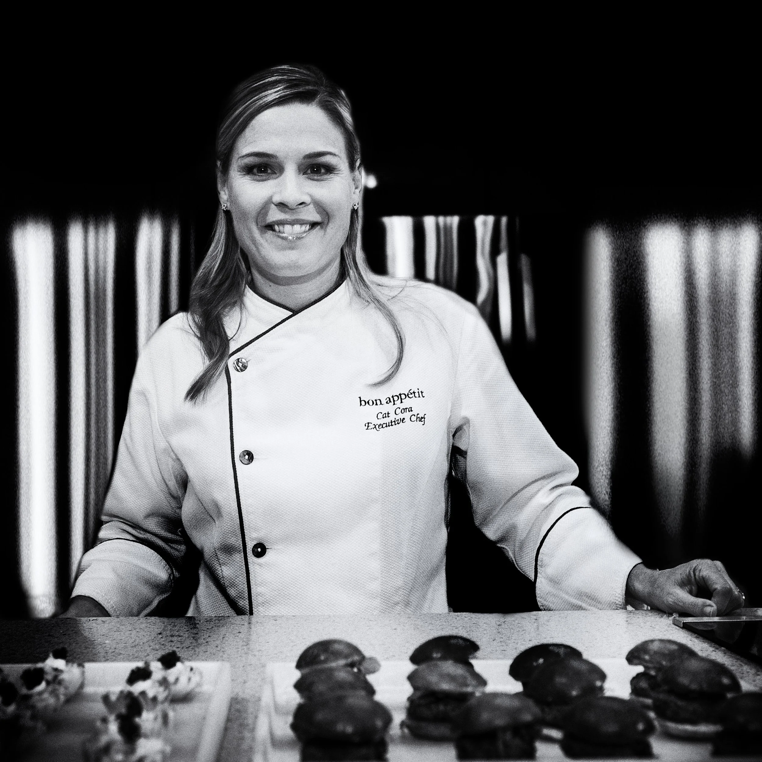 A smiling female chef in a white chef's coat standing behind a counter with baked goods, including cookies or pastries, on display.