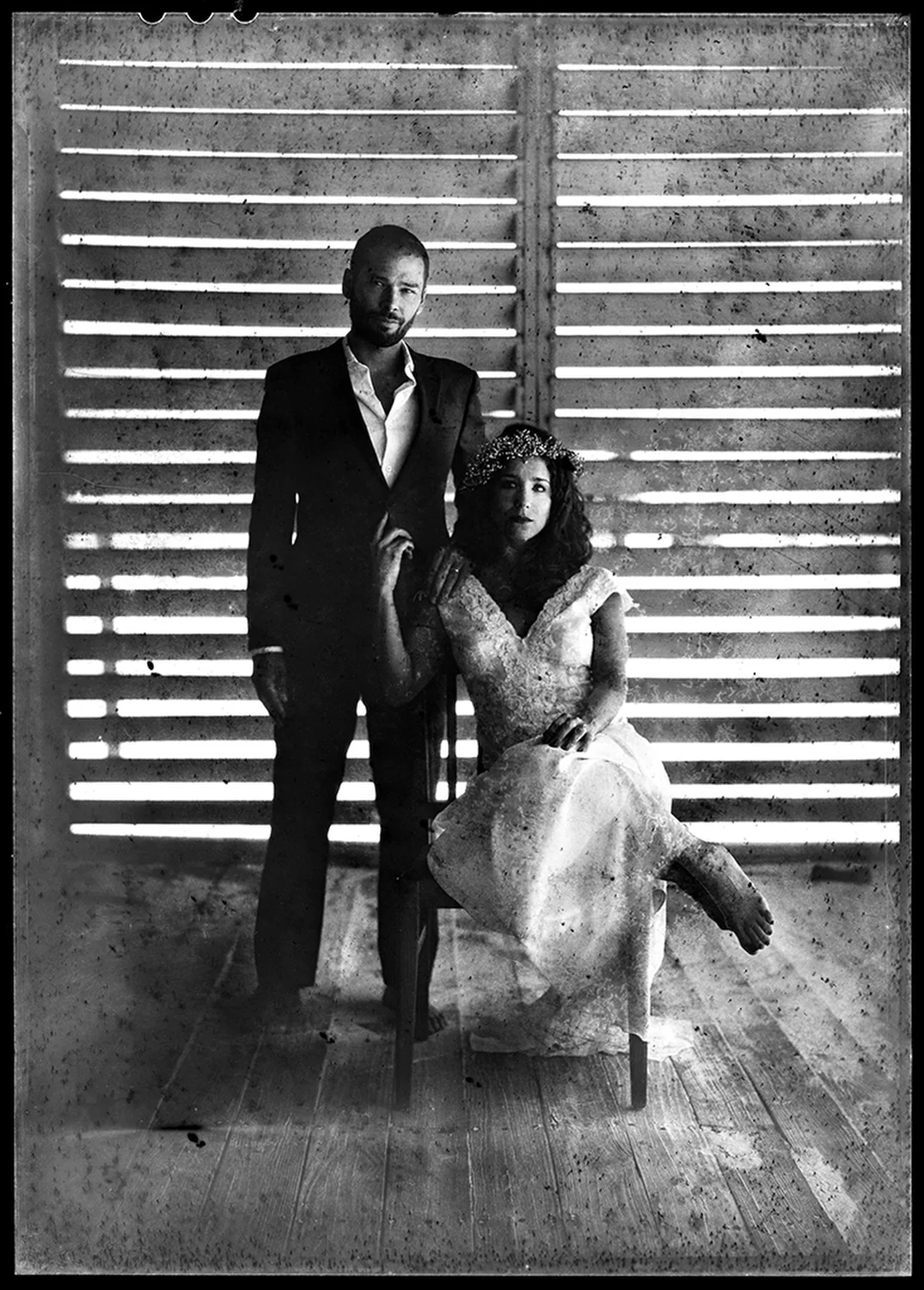 A black and white photo of a man in a suit standing next to a seated woman in a lace dress and floral headpiece, with a wooden wall background with horizontal slats.