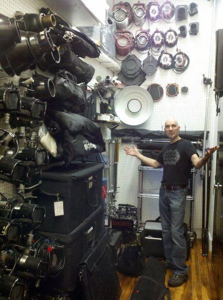 A man in a retail store or equipment shop standing near a wall of stage or film lighting equipment and accessories, with some cameras and a large speaker on display.
