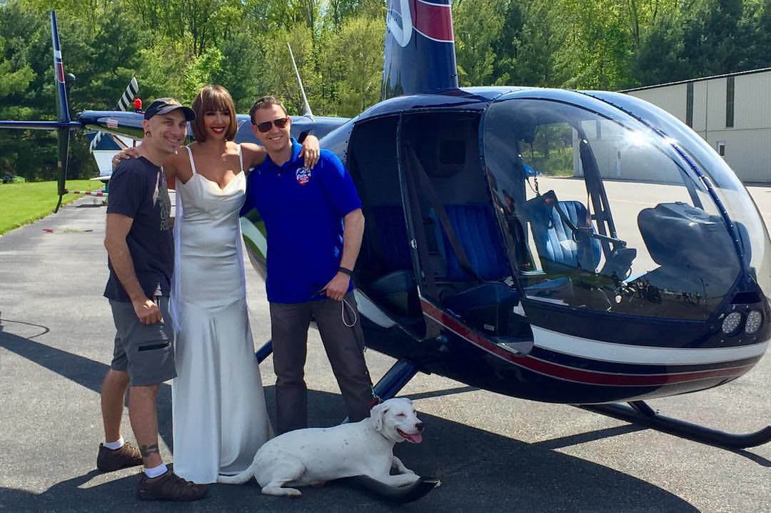 Three people standing in front of a helicopter with a dog sitting on the ground nearby.