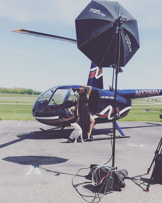 A woman in a black dress standing next to a white dog and a black helicopter on an airstrip. A large black umbrella is set up for shade, and there are bags and equipment on the ground.