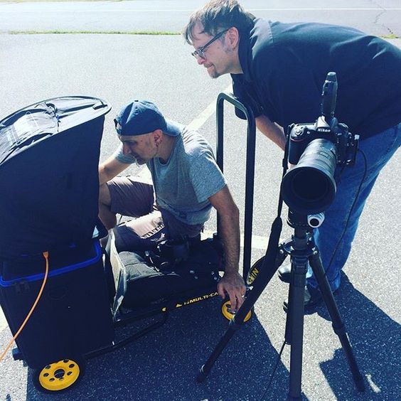 Two men setting up camera equipment on a paved outdoor surface, one adjusting the camera and another leaning over a cart.