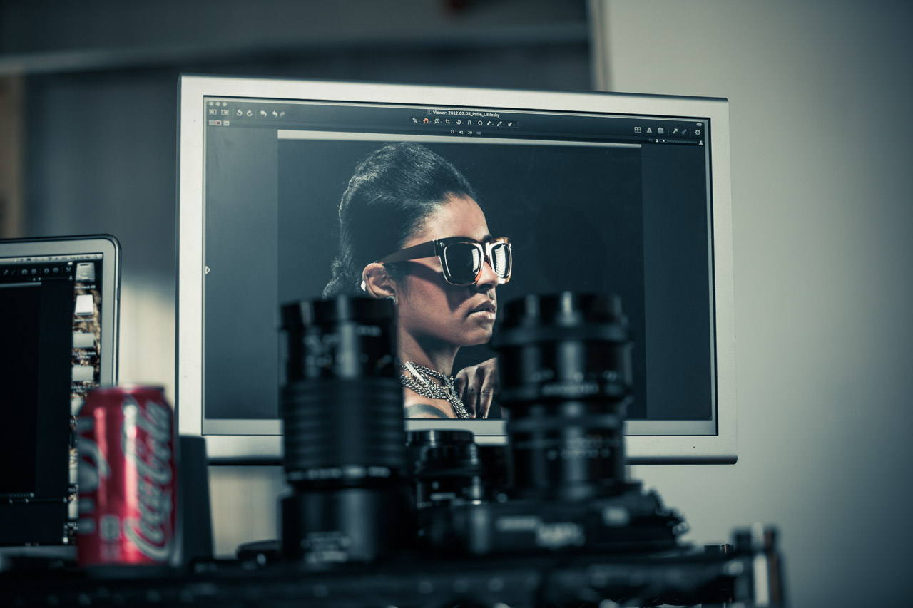 A computer monitor displaying a fashion photograph of a woman wearing sunglasses and jewelry, with camera lenses and a soda can in the foreground.