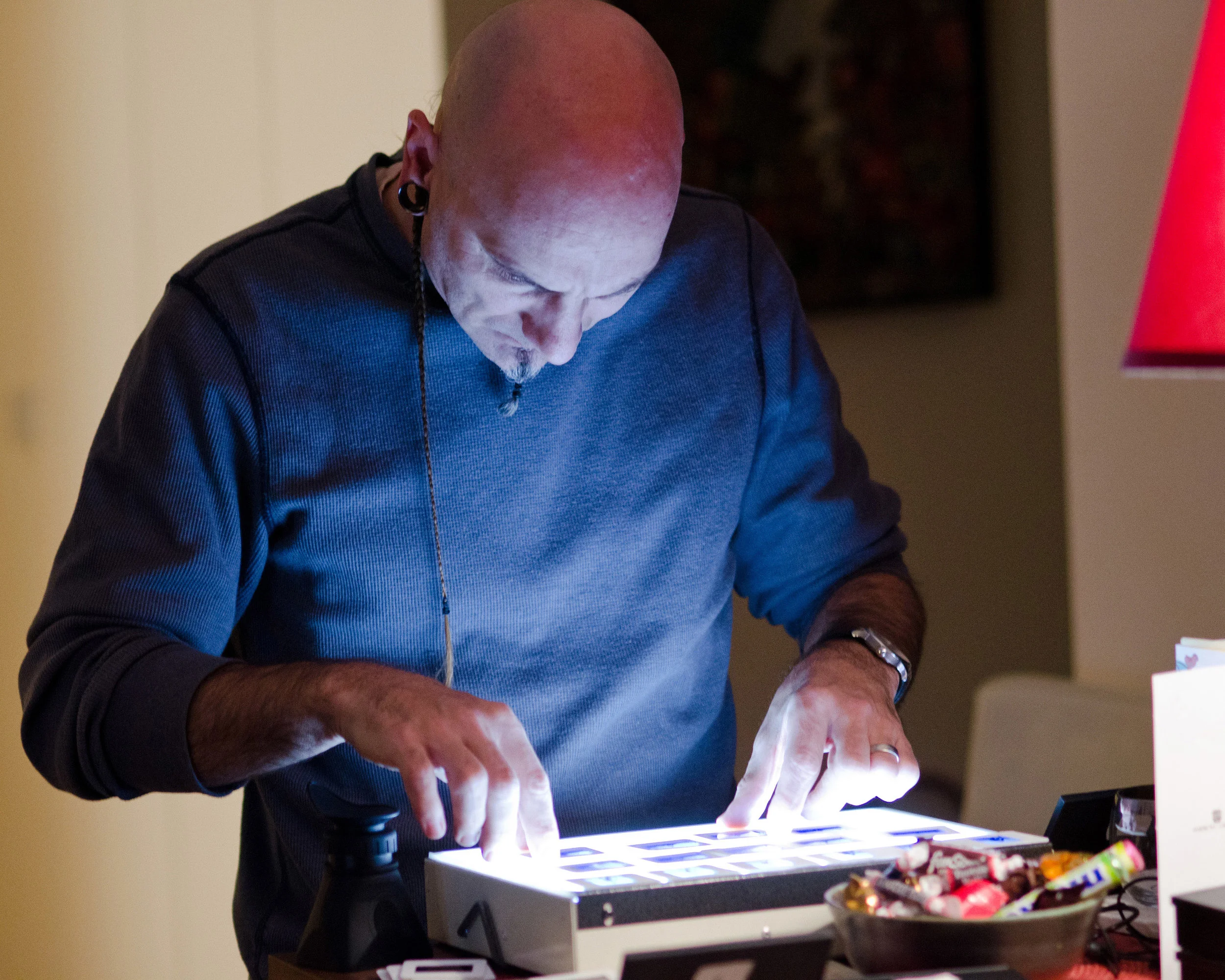A bald man with earrings and a goatee, wearing a blue shirt, looking down and interacting with a lighted electronic device on a table.