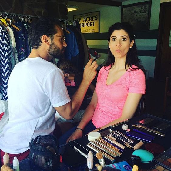A woman with long dark hair wearing a pink T-shirt sitting at a makeup station, having makeup applied, with makeup brushes and tools on the table. A man with glasses and a white T-shirt is standing next to her, holding a makeup brush or tool.