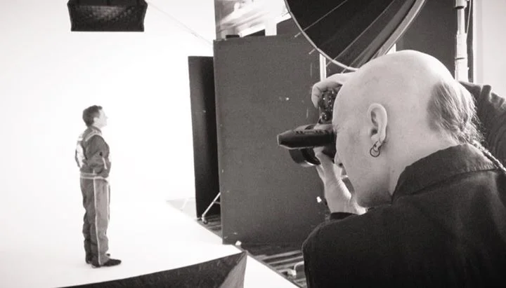 Photographer taking a picture of a young boy standing on a white studio background.