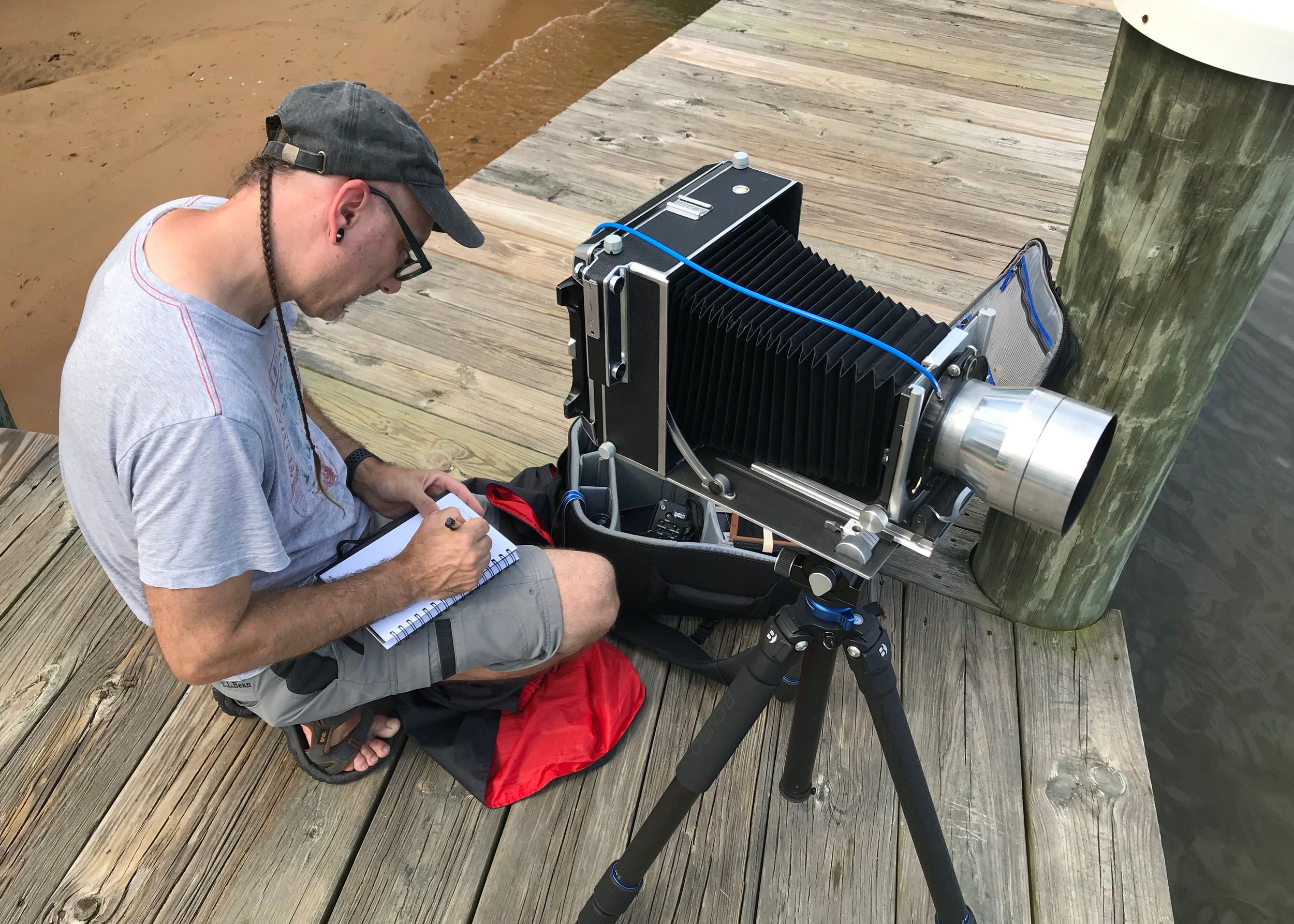 A man with glasses and a cap inspecting a large camera on a tripod on a wooden pier.