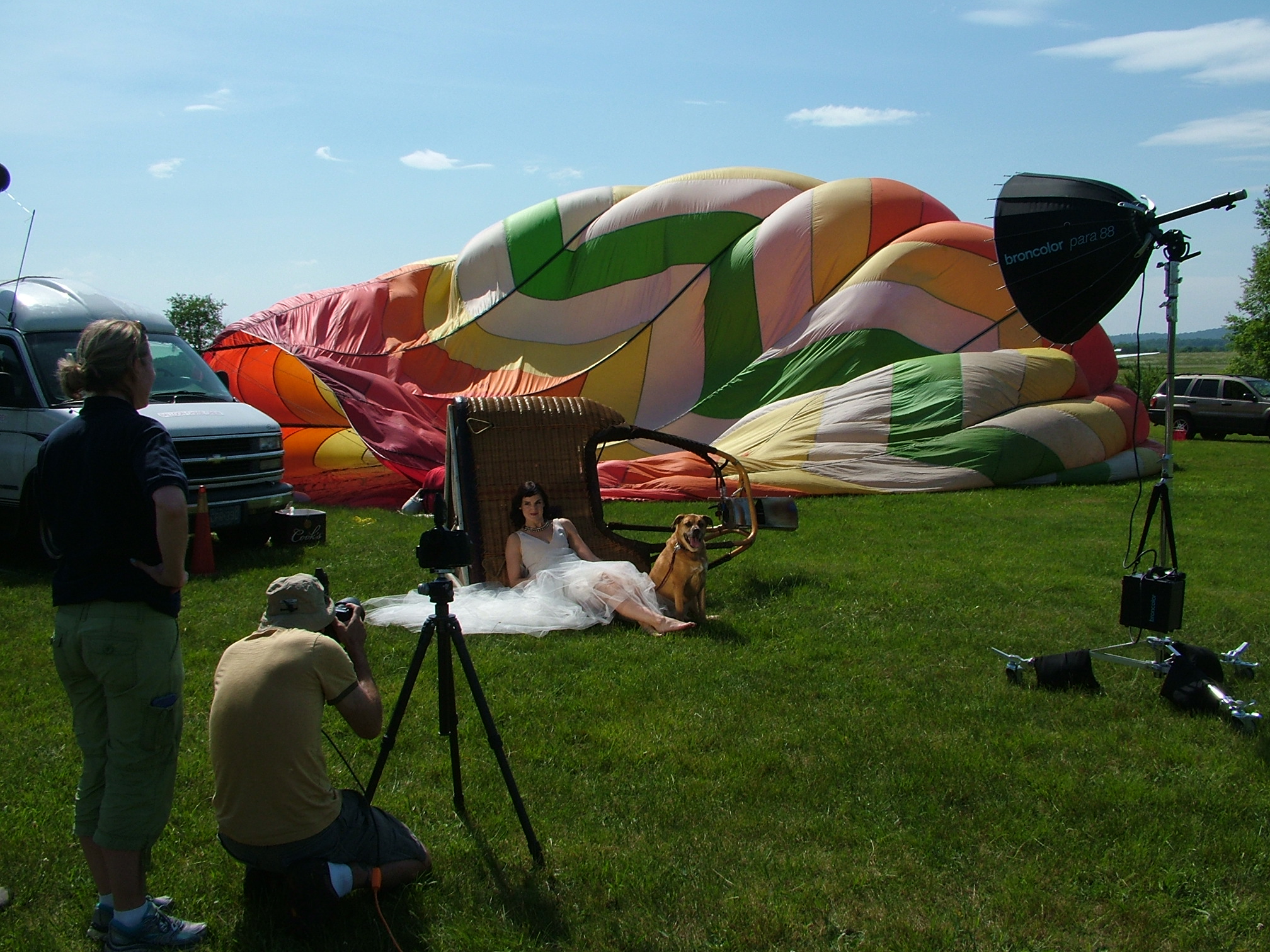 A photo shoot scene of a woman in a wedding dress with a dog, set outdoors near a balloon being prepared for inflation, with photography equipment and people filming.