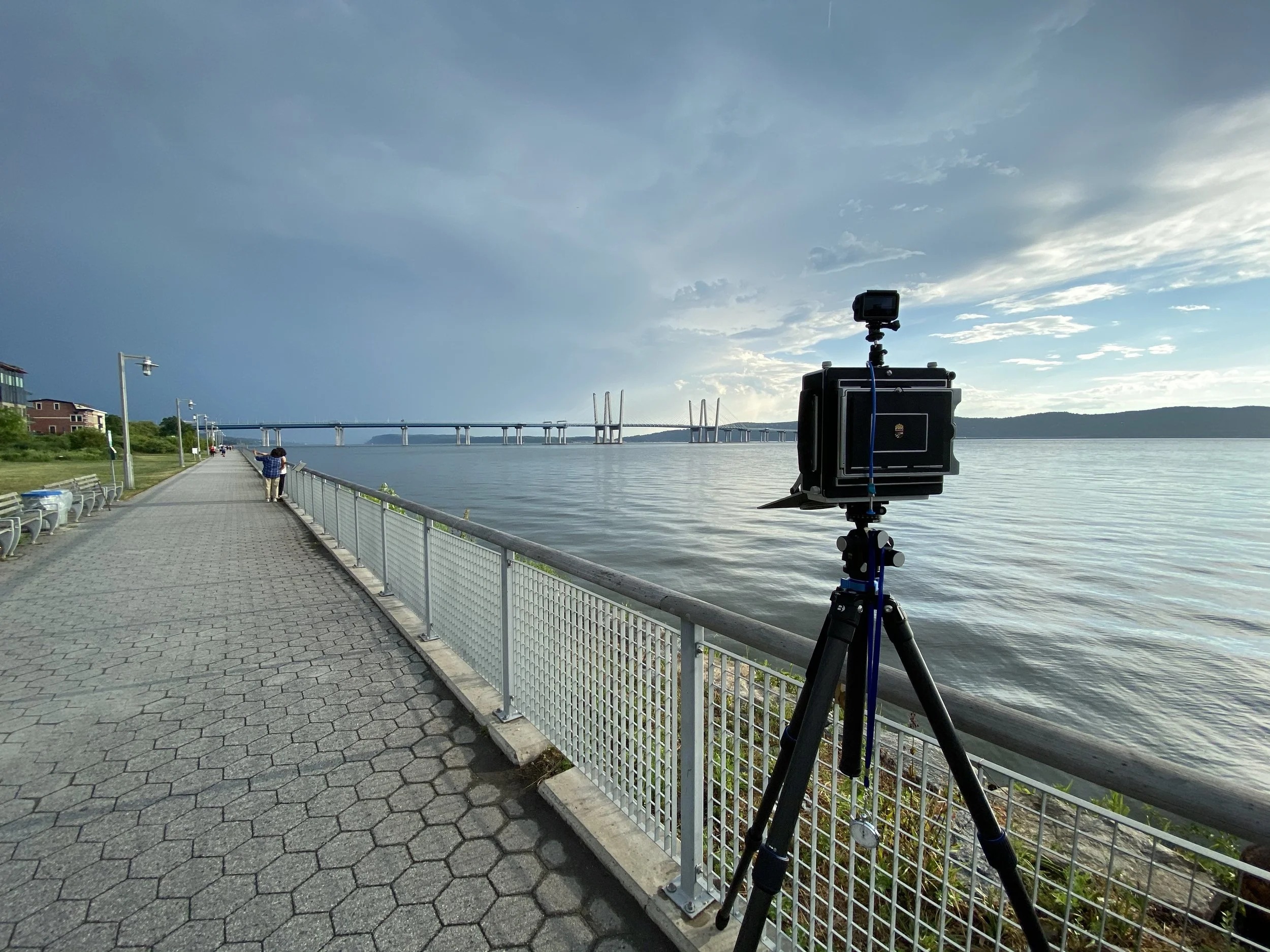 A camera on a tripod facing a waterfront promenade with a bridge in the background, trees, and a partly cloudy sky.