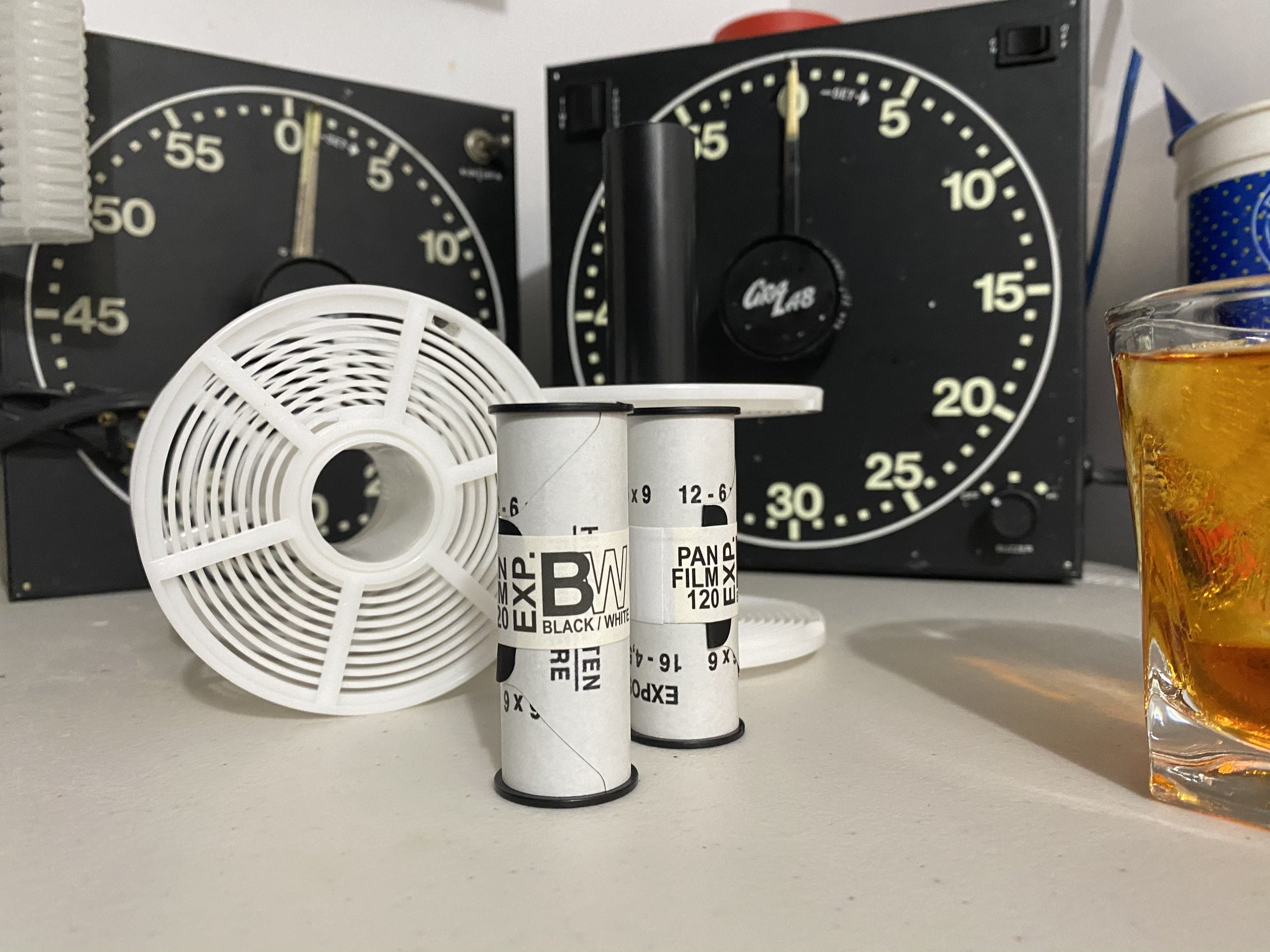 Two vintage timers, a white plastic fan, and a glass of amber-colored liquid on a white surface. In the background are two large black and white chess clocks.