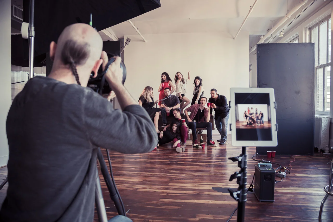Photographer taking a group photo of young people in a photography studio with a white wall background, some sitting, standing, and posing.