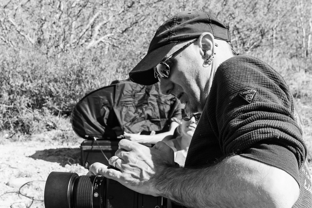 A man wearing a baseball cap and sunglasses is looking at a camera in an outdoor setting with trees and bushes in the background.